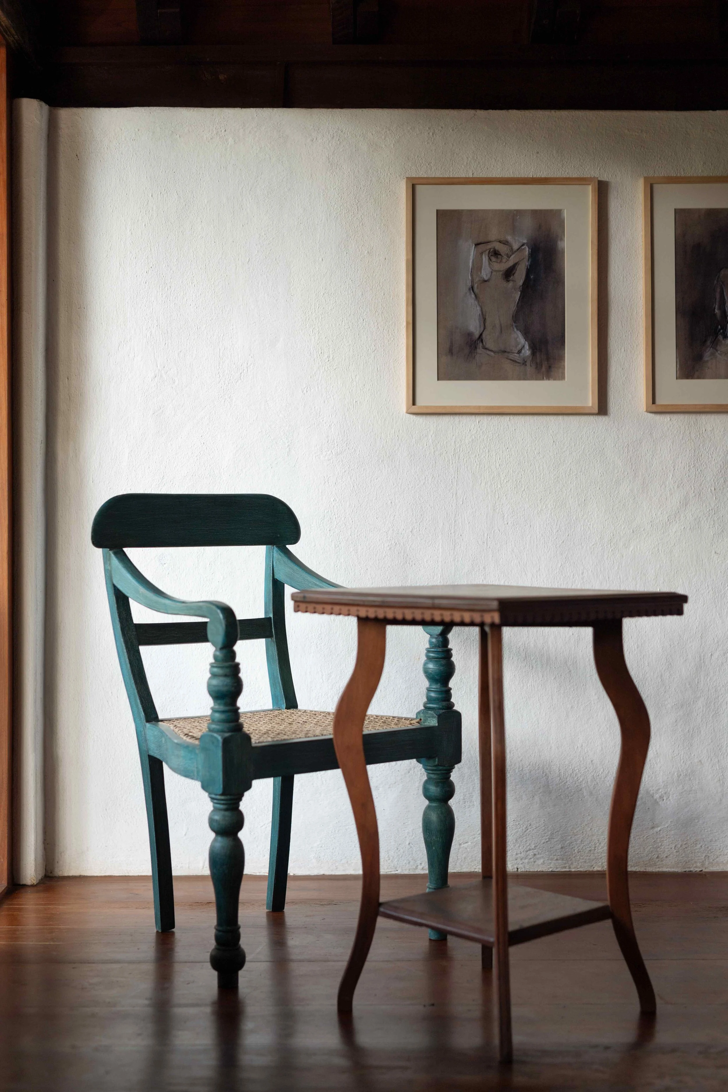 Interior scene with a wooden chair featuring a spotted woven seat and a small decorative table with curved legs, set against a white wall with framed abstract art.