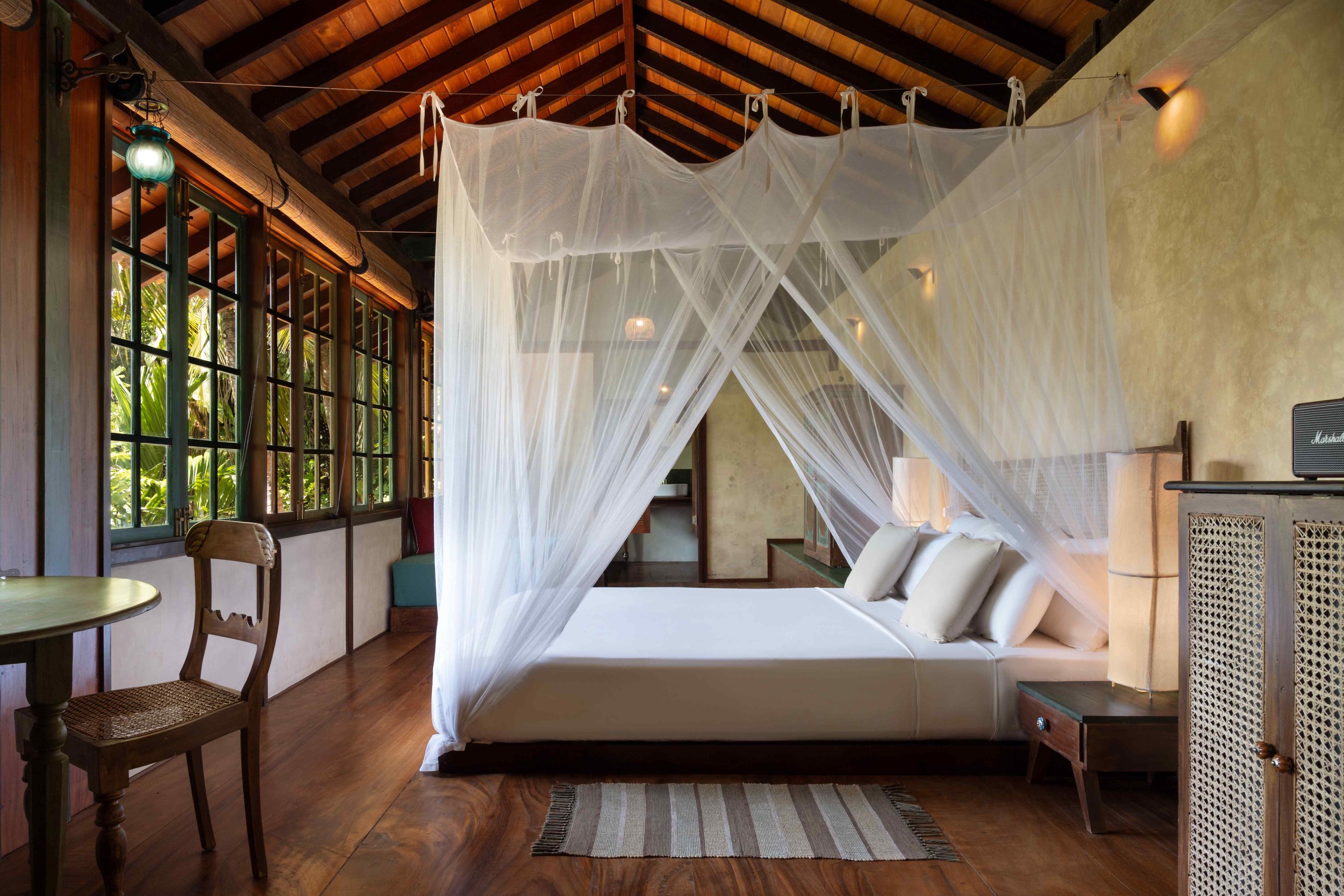 A bedroom with a canopy bed, white bedding, and a mosquito net. The room has wooden floors, large windows, and rustic furniture, with sunlight coming through the windows.