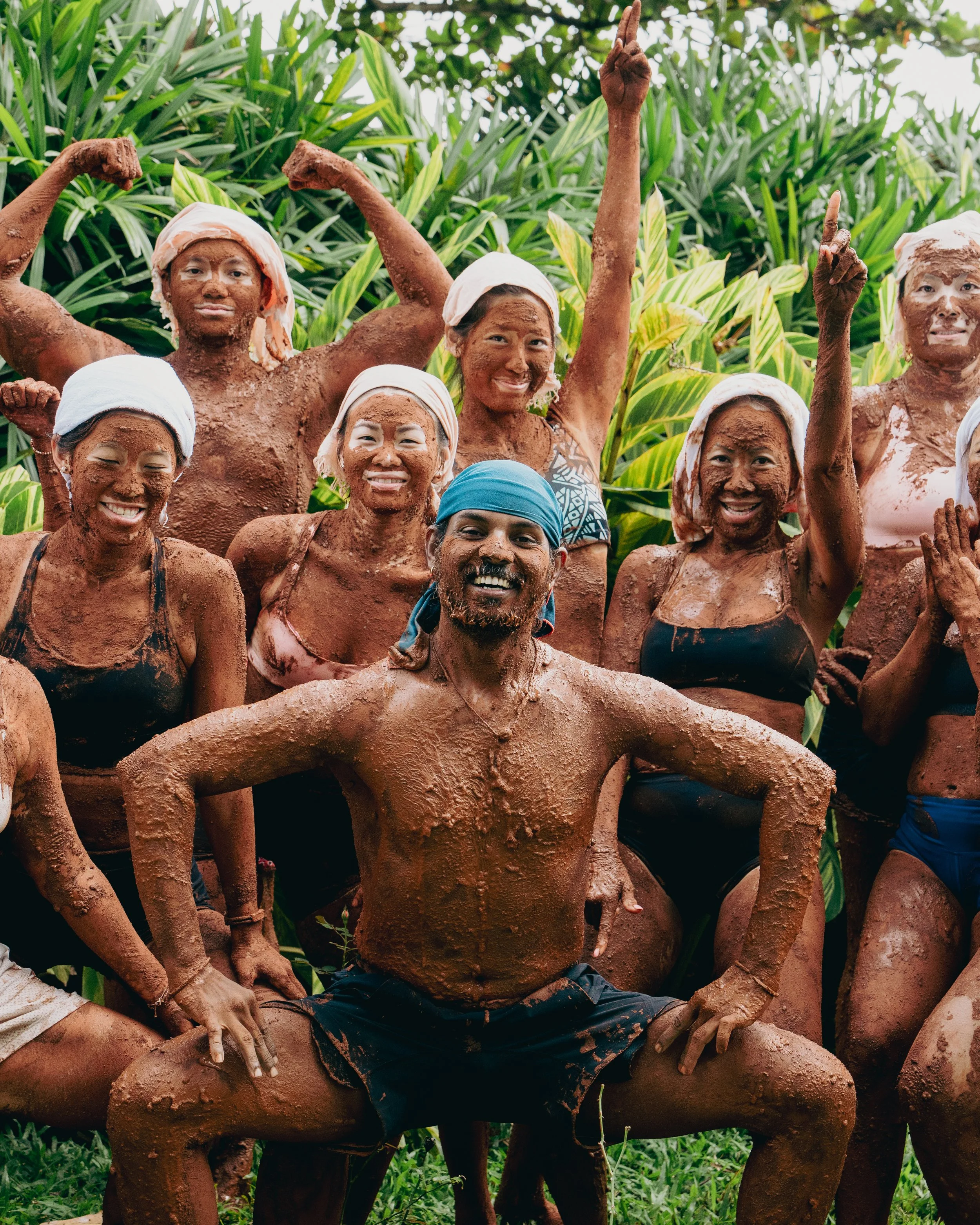 A group of people covered in mud, posing and smiling among lush green plants, some with arms raised.