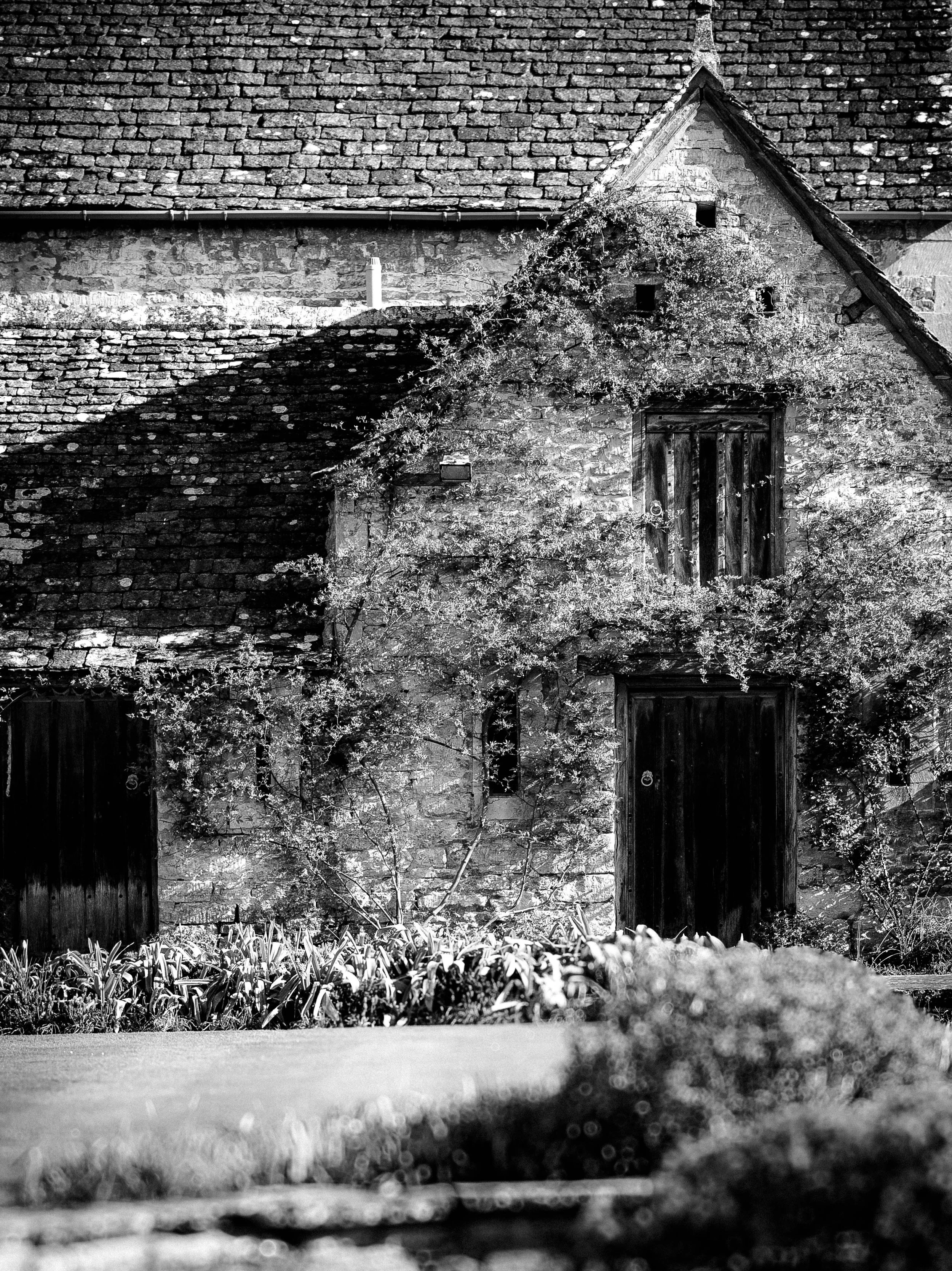 A black and white photo of an old Cotswold stone house with wooden doors, partially covered in climbing plants, with a tiled roof and a small garden in the foreground.