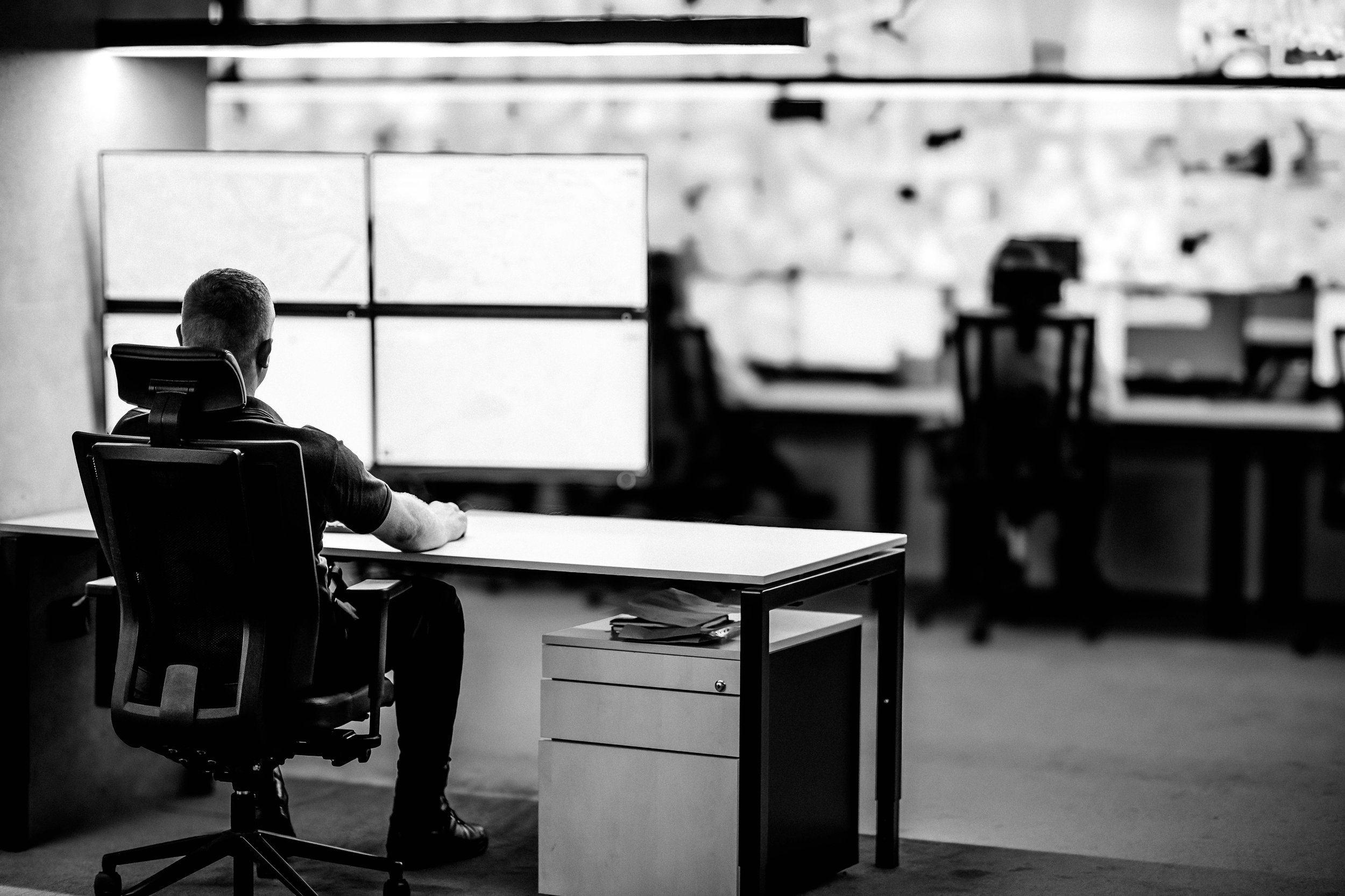 A man sitting at a desk in a Security Operations Centre, facing four computer monitors in a 2x2 configuration, with a blurred background and a filing cabinet nearby.