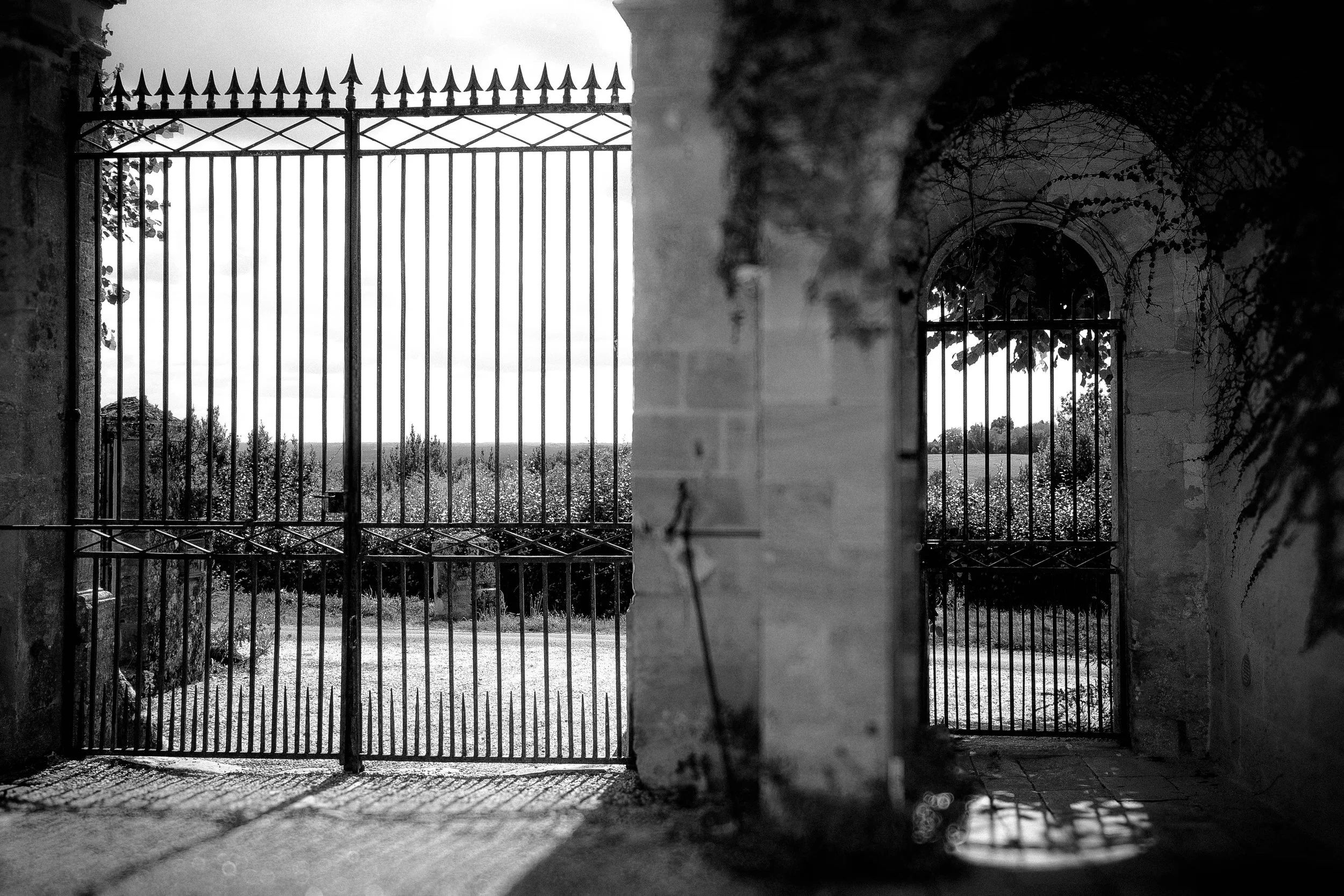 Black and white photo of metal gates with vertical bars and pointed tops, within stone archways, casting shadows on the ground, overlooking a field with trees in the distance.