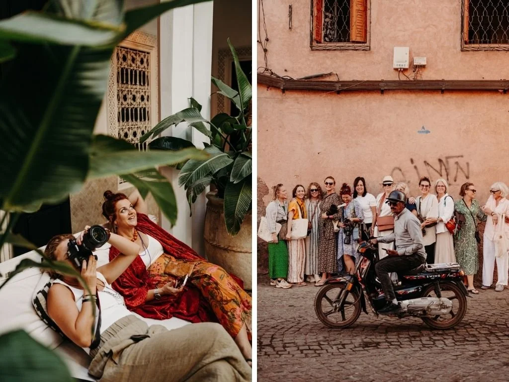 A woman with red hair sits on a bed, smiling, with a photographer taking her picture. She is surrounded by green plants in a cozy, decorated indoor space. Outside, a group of people are lined up on a cobblestone street, with a man riding a scooter past them; the scene appears to be in an urban area with an old, pinkish building in the background.