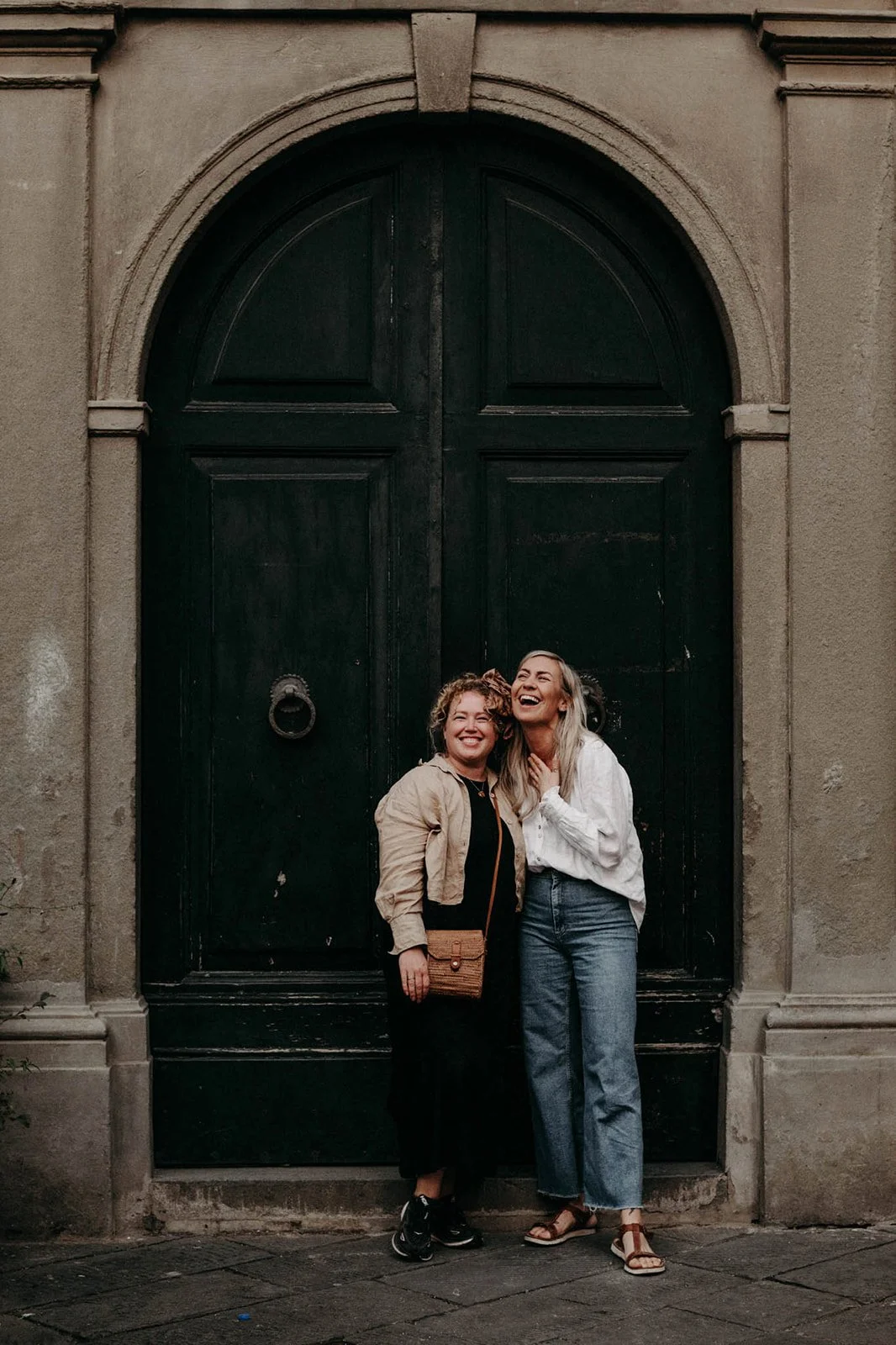 Two women standing in front of a large black arched door, smiling and laughing together.