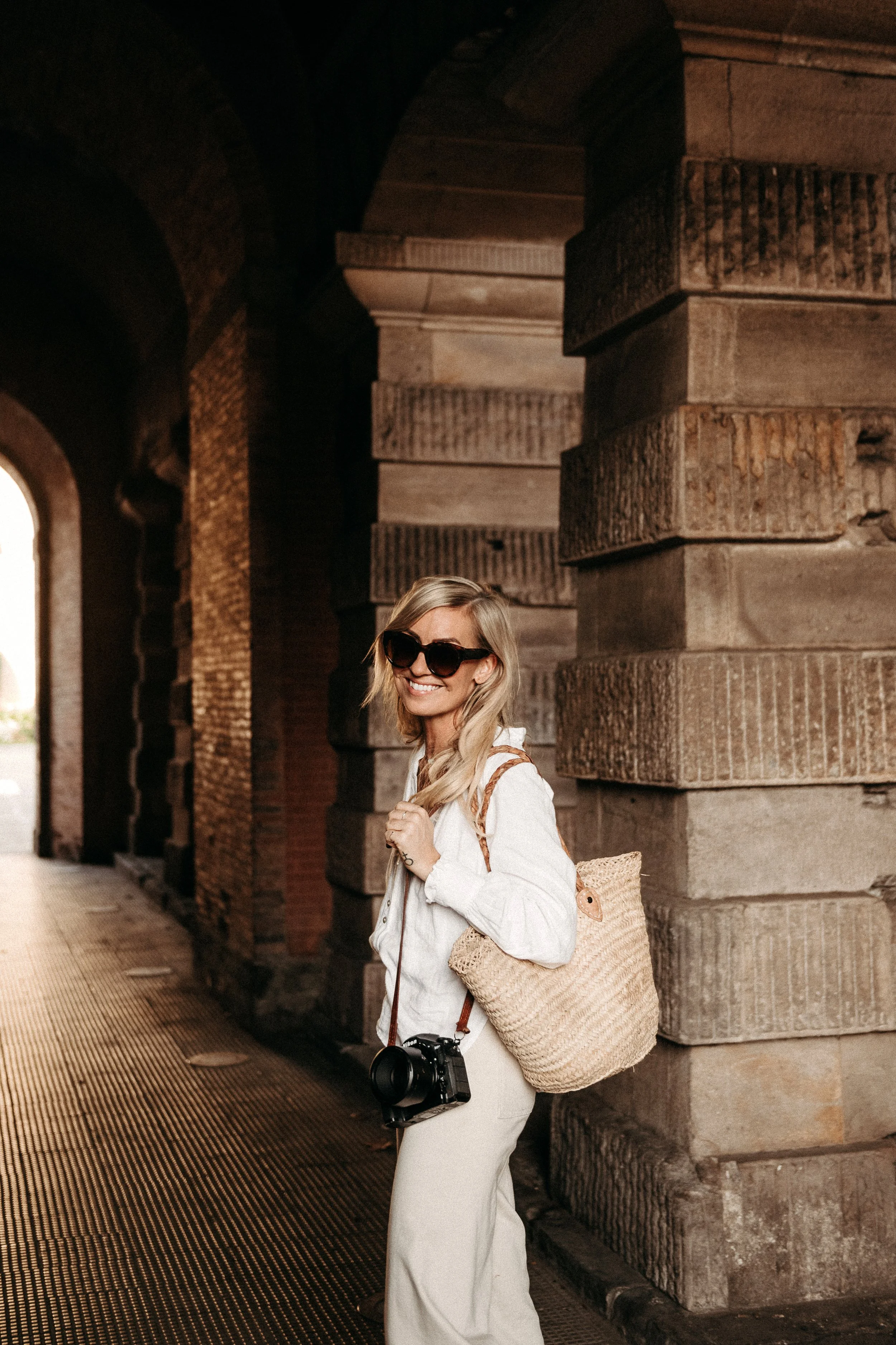 A woman wearing sunglasses, a white outfit, and carrying a camera around her neck, stands under an arched stone bridge with large brick columns, smiling.
