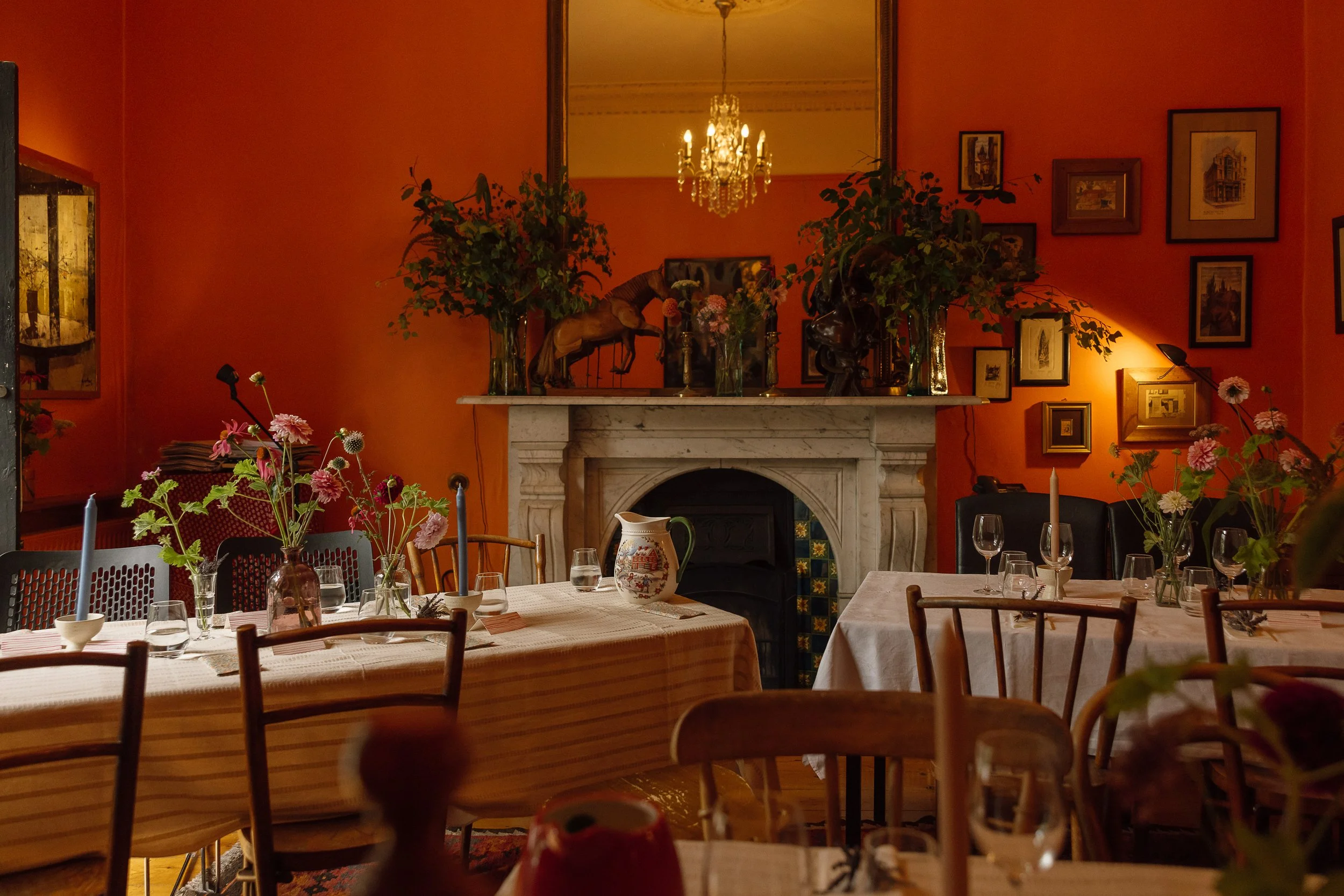 A warmly lit dining room with a marble fireplace, several tables with floral centerpieces, and a chandelier reflected in a mirror above the fireplace. The walls are decorated with framed artwork.