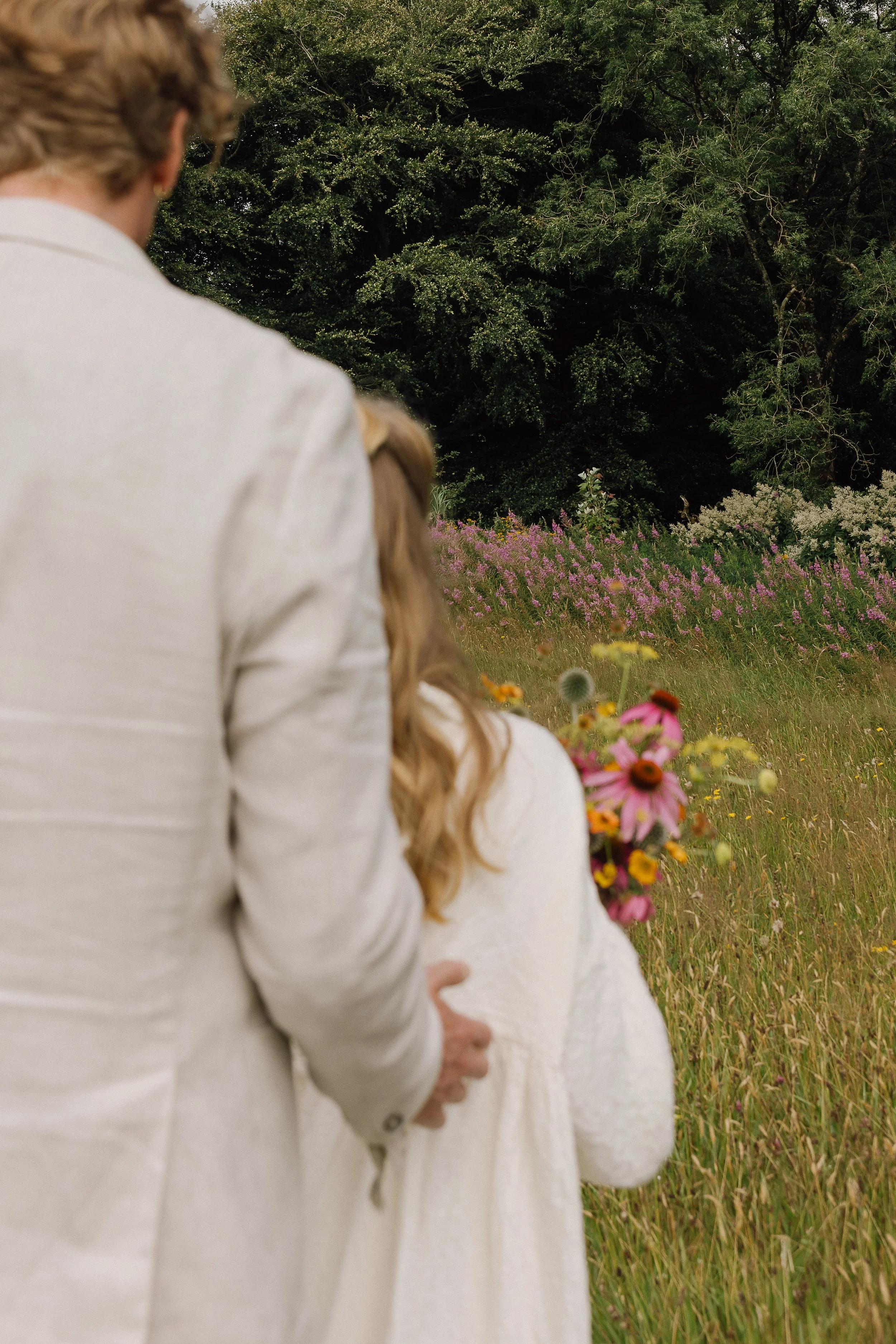 A woman dressed in an elegant white outfit holding a colorful bouquet of flowers, standing in a lush green field with tall grass and pink wildflowers, surrounded by trees in the background.