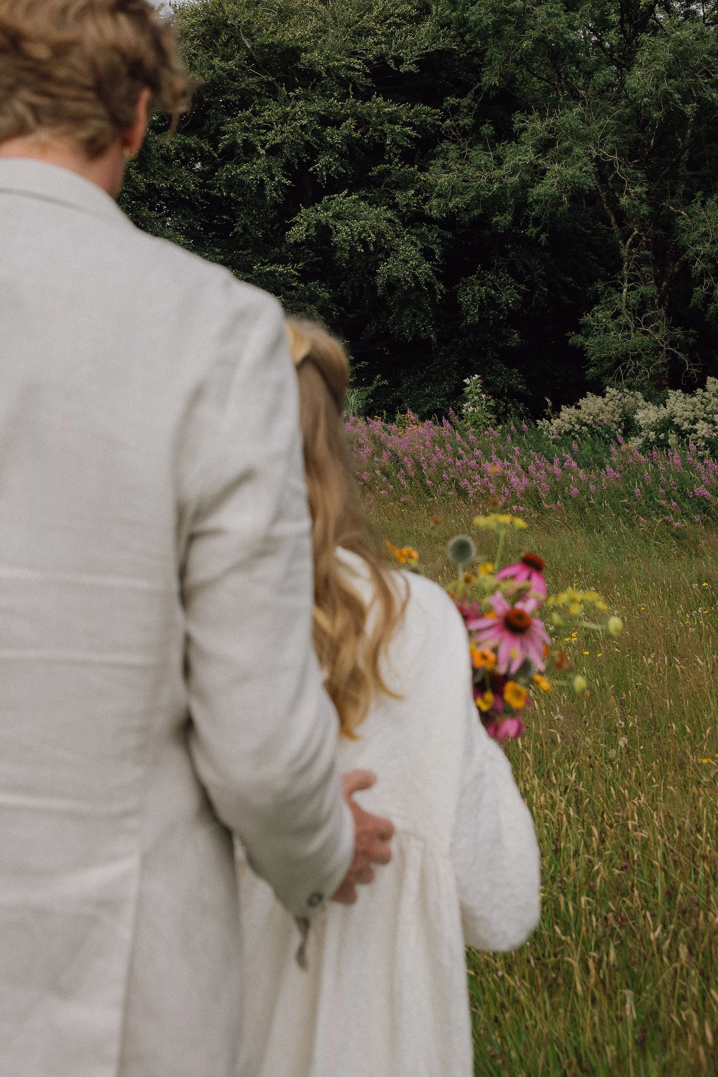 A man and a young girl standing in a field of wildflowers, holding hands, with trees and colorful flowers in the background.