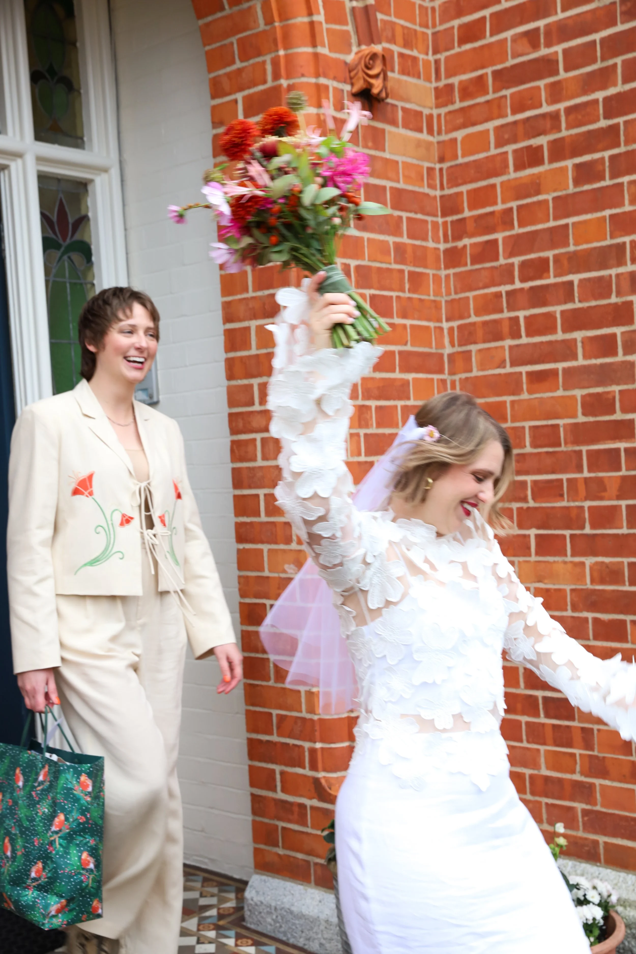 A bride in a white wedding dress and veil is joyfully raising a bouquet of colorful flowers, while a woman in cream-colored pants and jacket with floral embroidery smiles nearby, holding a gift bag, in front of a brick wall.