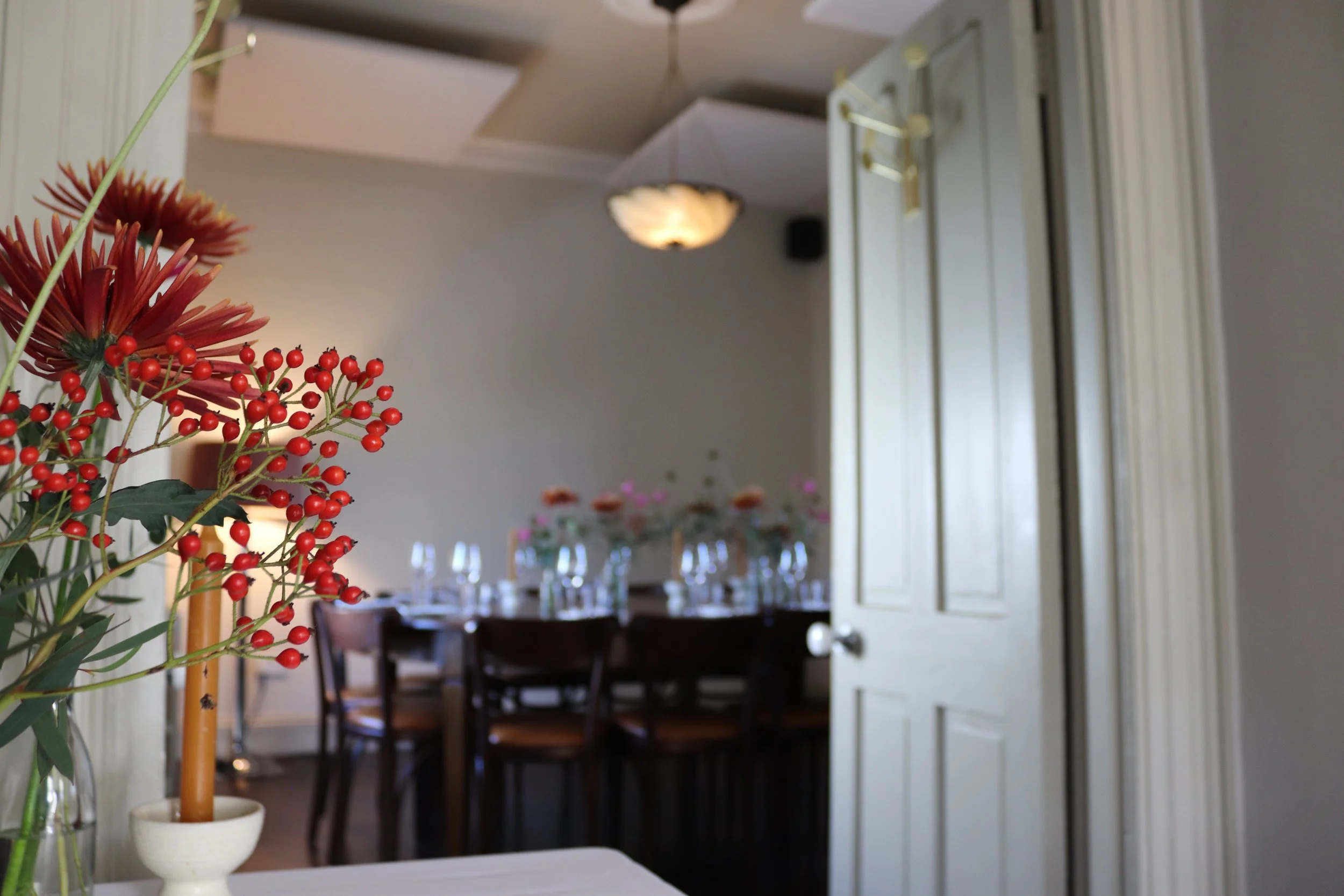 Interior of a dining room with a table set with wine glasses and floral centerpieces, partially seen through a doorway, with a red flower and berries in a vase in the foreground.