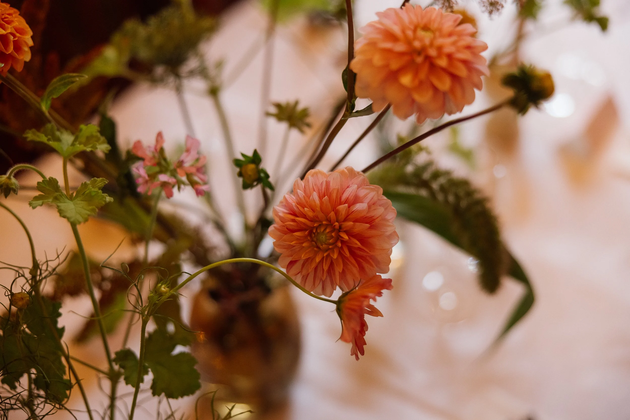 Close-up of a bouquet with pink and orange dahlias, green leaves, and small pink flowers, in a blurred background.