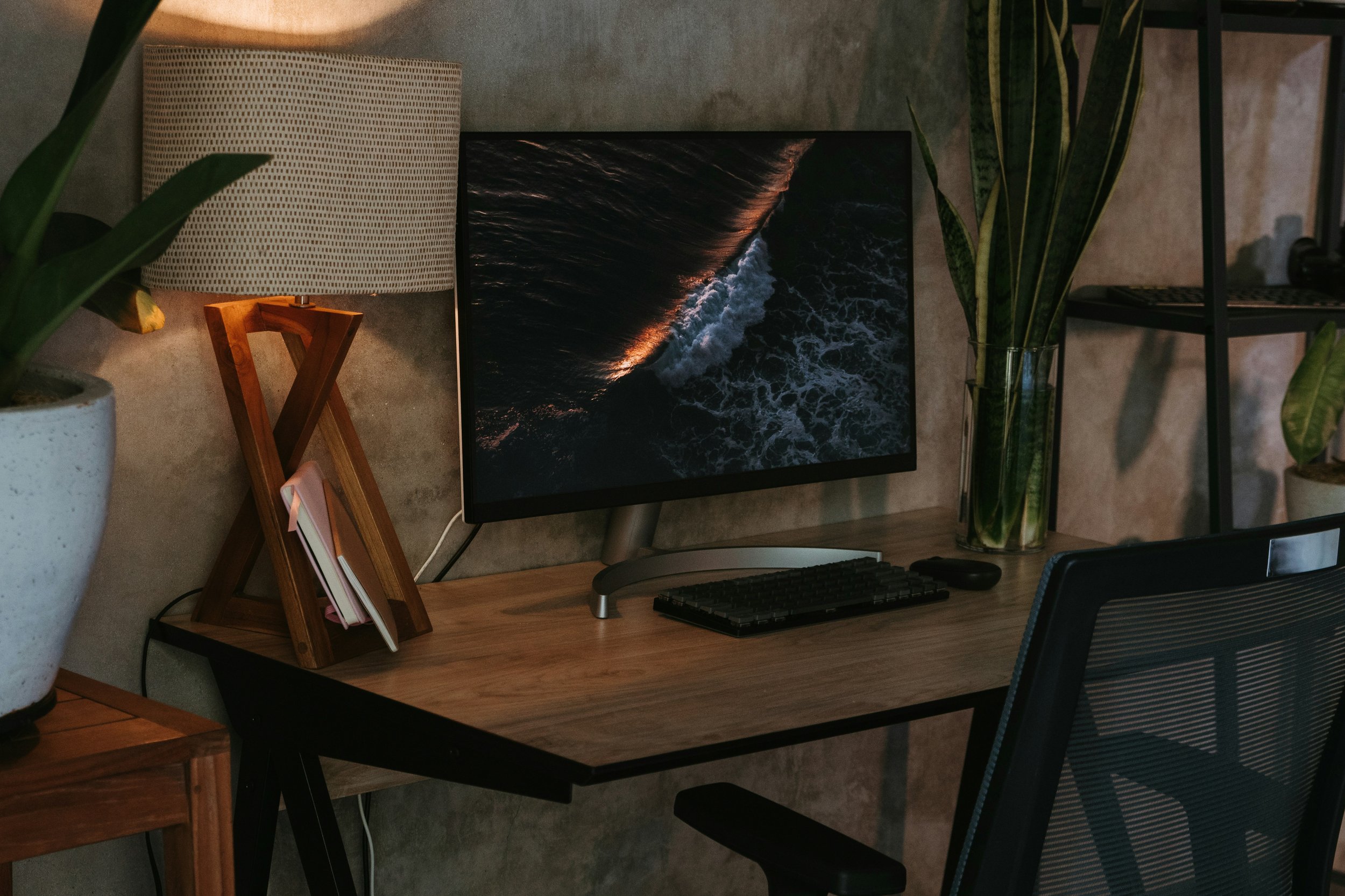 Home office setup with a computer monitor displaying a seascape image, wooden desk, lamp, keyboard, chair, plant, and decorative shelves.