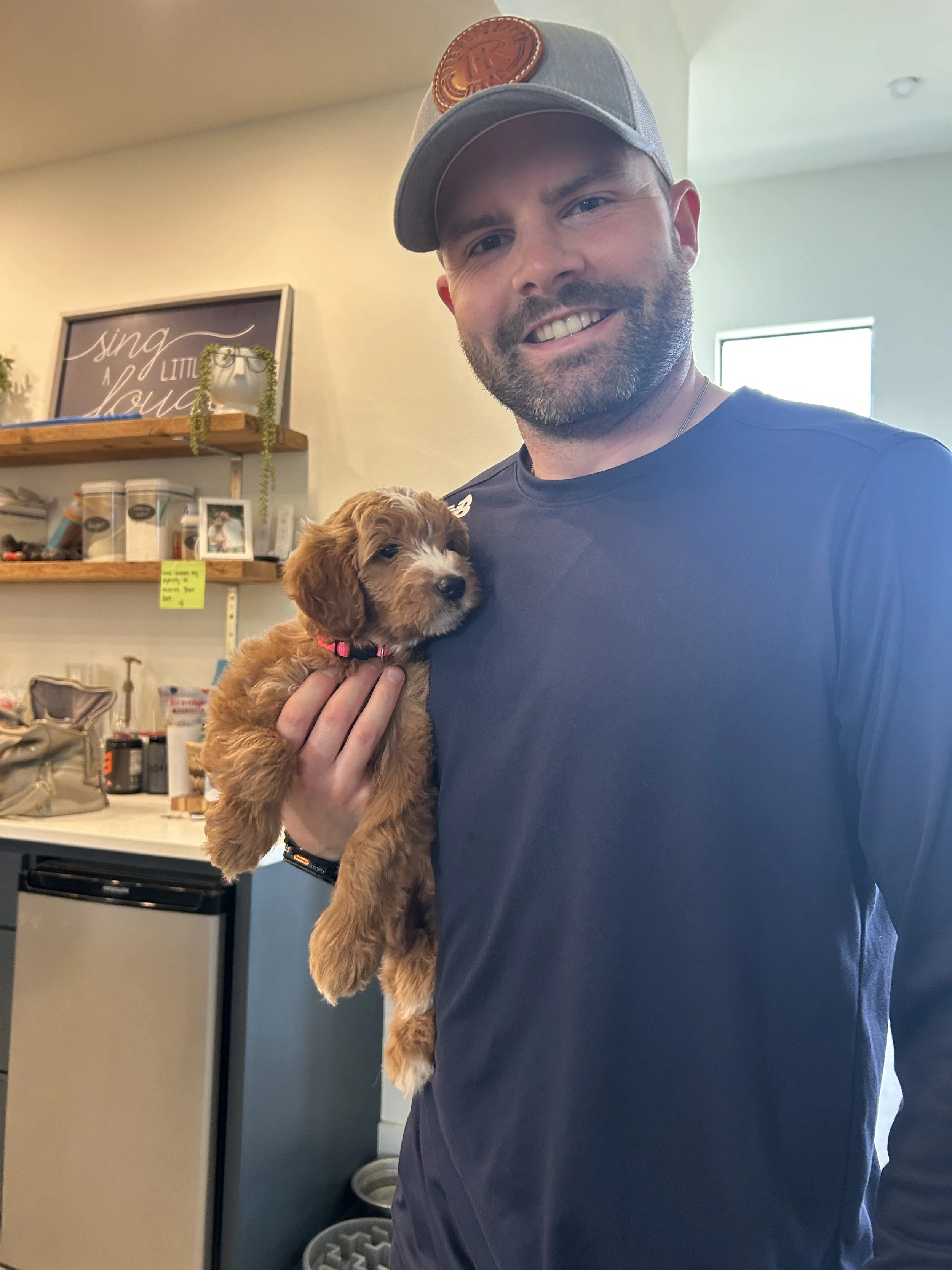 A man holding a small brown puppy indoors, smiling at the camera.