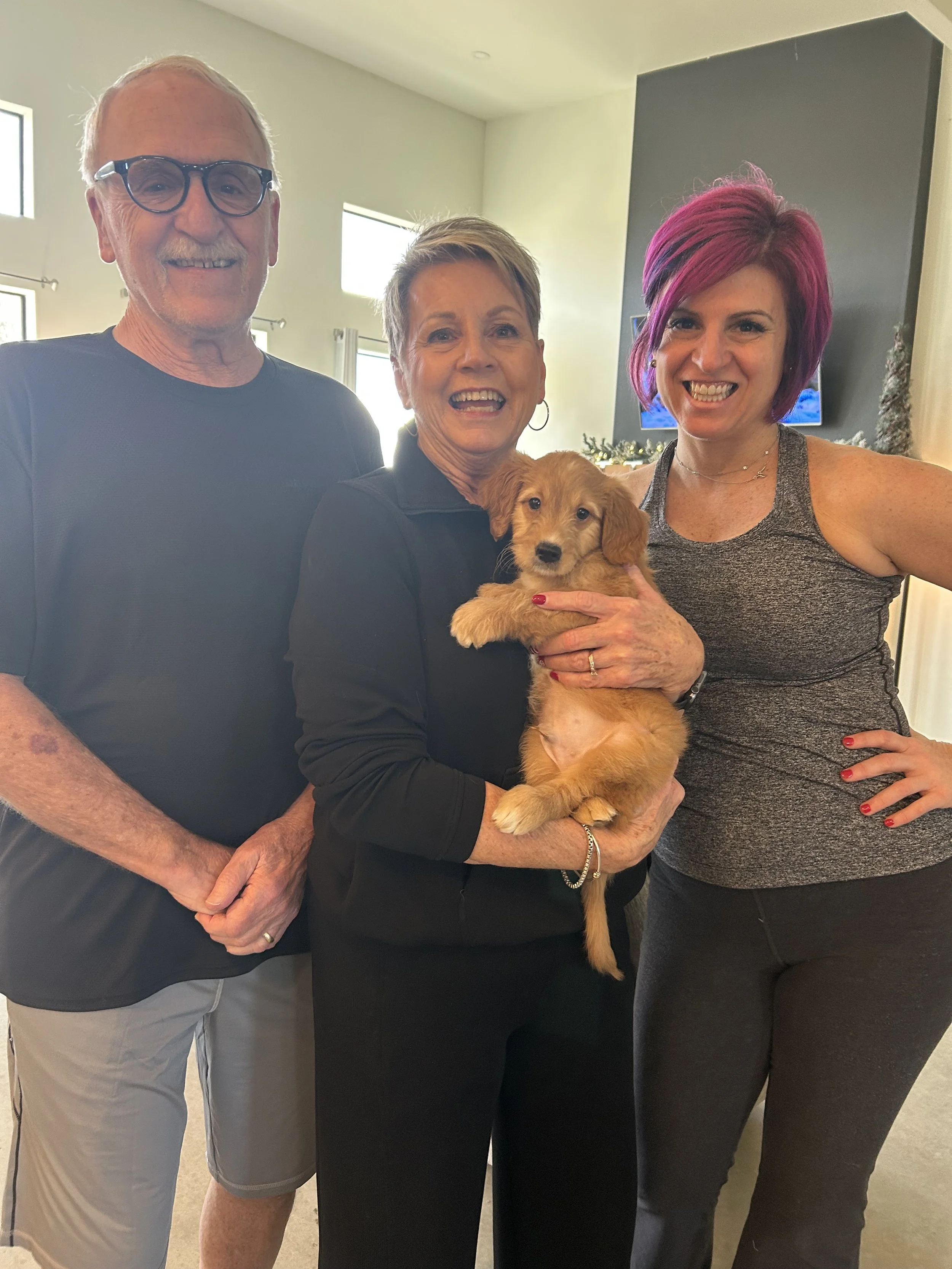 Three people and a puppy posing indoors with windows in the background. The woman in the middle is holding a small brown puppy, and the other two women are standing beside her.