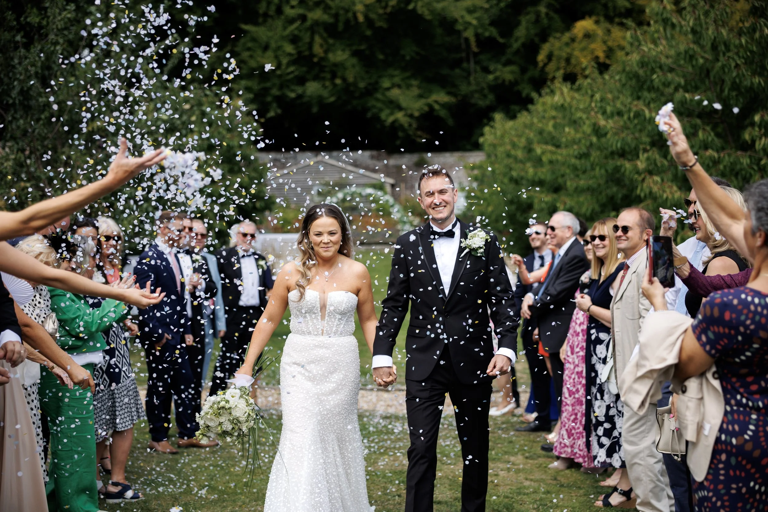 A newly married couple walks hand in hand through a crowd throwing confetti, smiling, outdoors with green trees in the background.