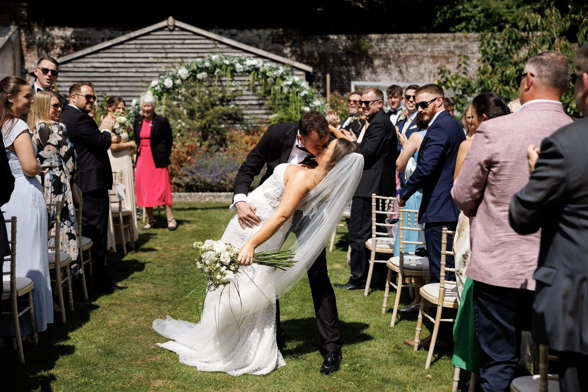 A bride and groom share a kiss during their outdoor wedding ceremony, surrounded by seated guests clapping and smiling.