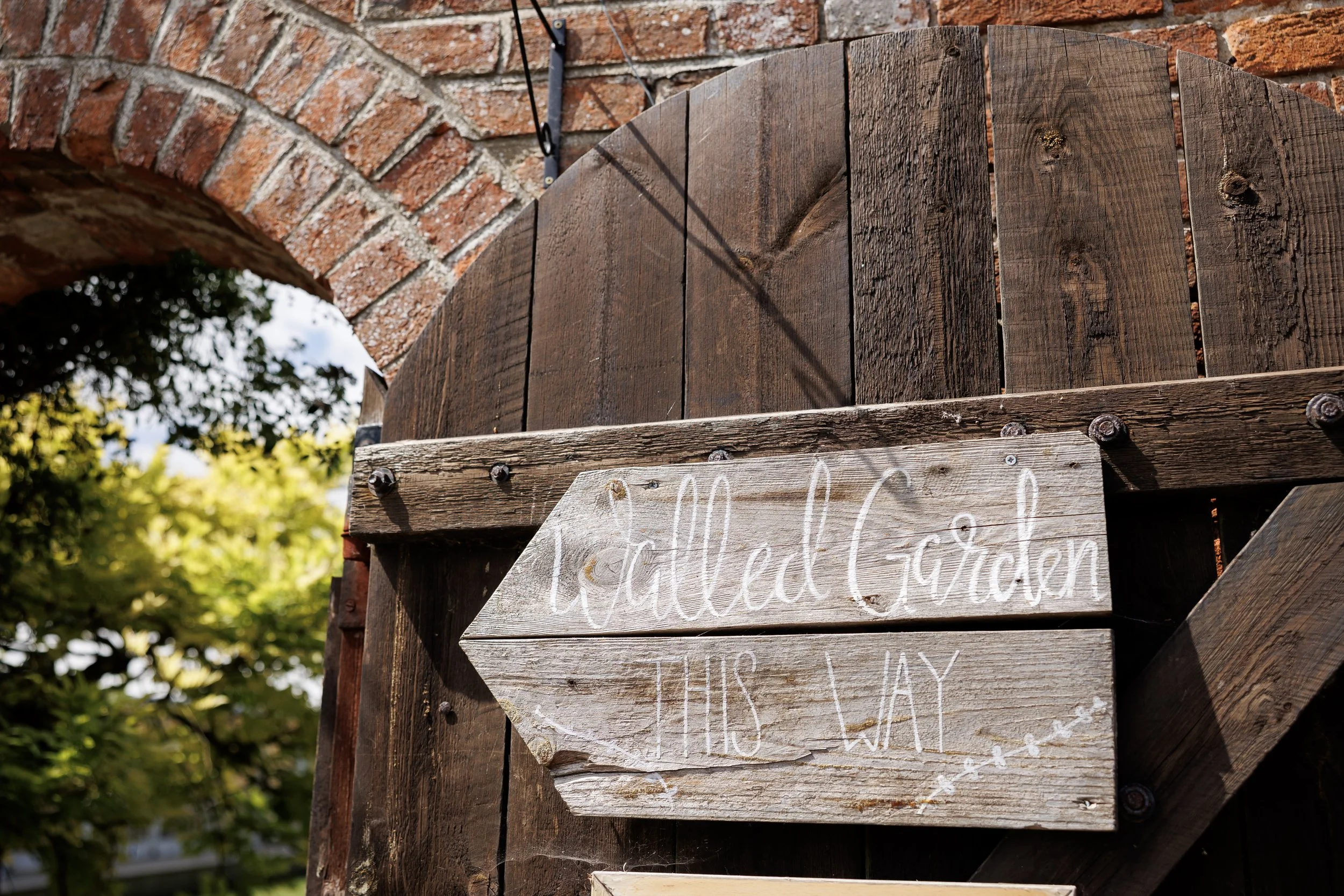 Wooden arrow-shaped sign reading "Walled Garden This Way" attached to a dark wooden gate with a background of brick and green trees.