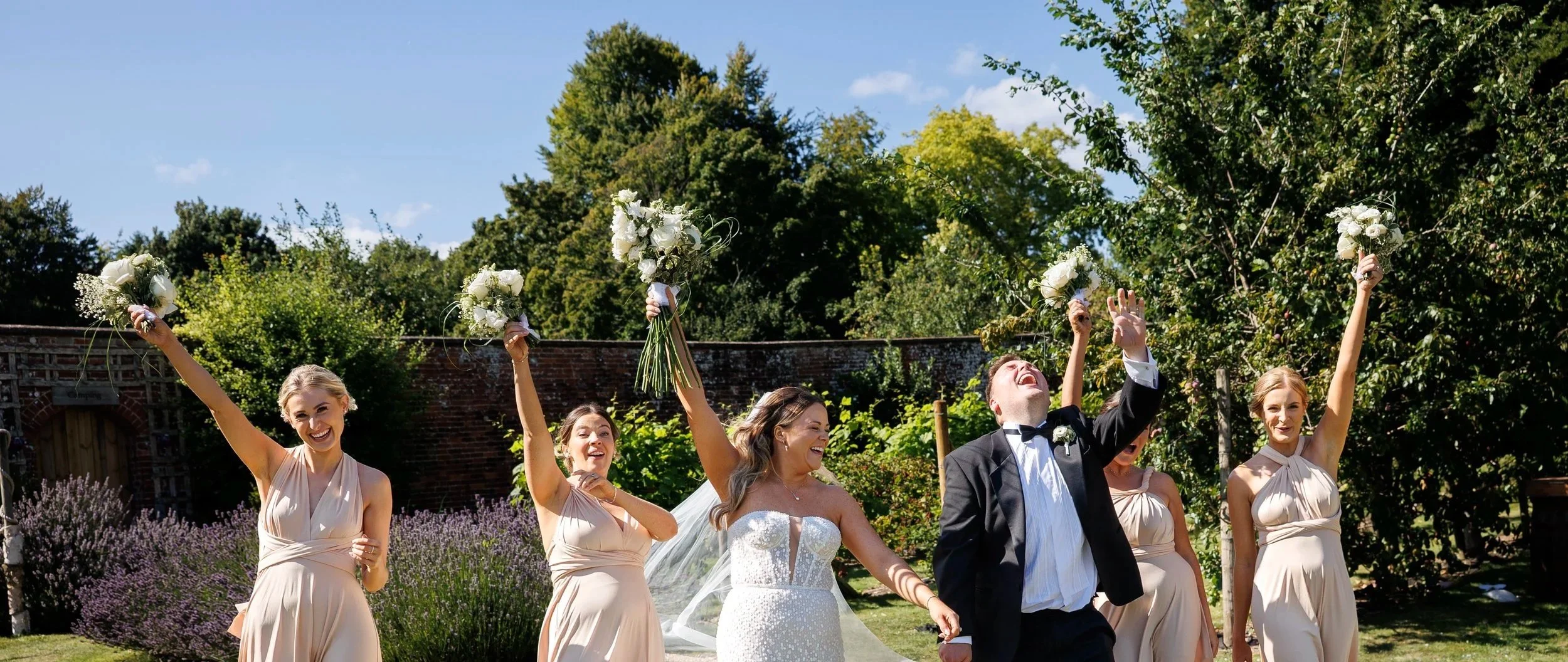 Wedding celebration outdoors with a bride, groom, and bridesmaids holding bouquets, smiling, and enjoying a sunny day in a garden.