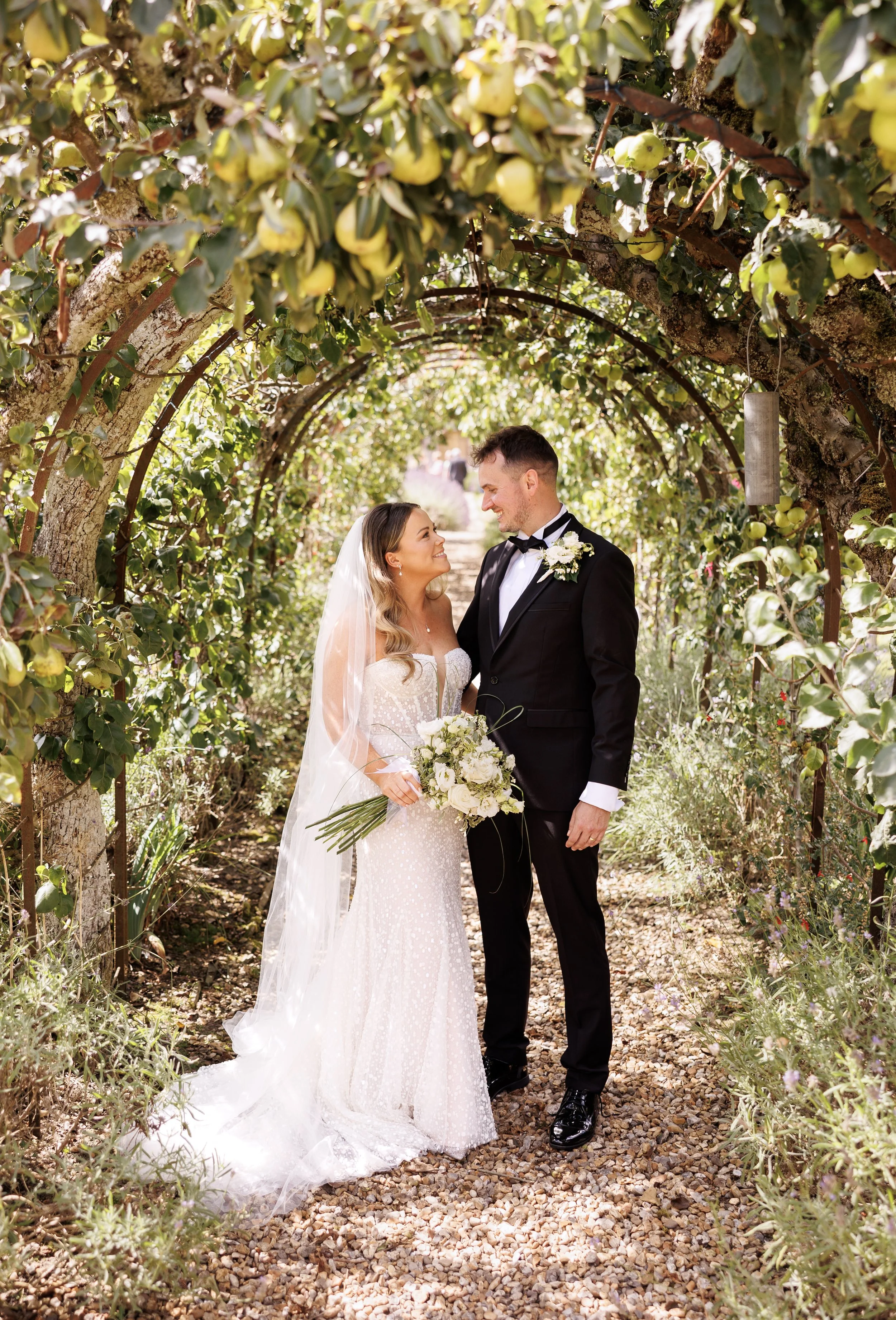 A bride and groom standing under a lush, leafy archway, gazing at each other, with the bride holding a bouquet of white flowers.