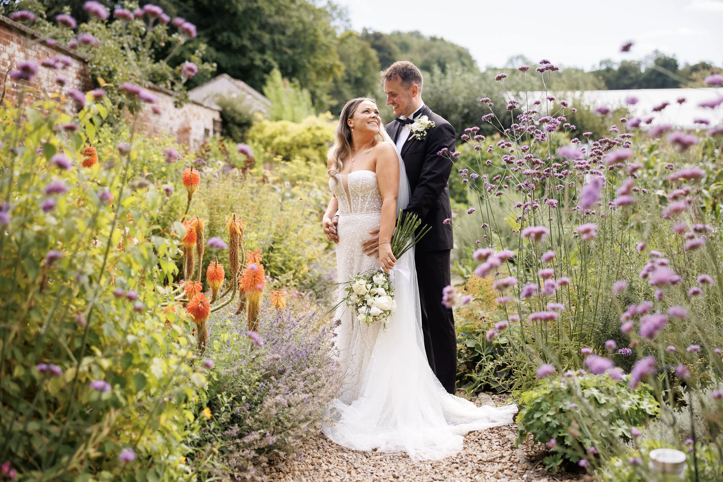 A newlywed couple stands together on a garden path, surrounded by colorful flowers. The bride wears a strapless wedding gown and holds a bouquet of white flowers, while the groom is dressed in a black tuxedo with a bow tie. They look at each other lovingly, smiling in a lush outdoor setting.