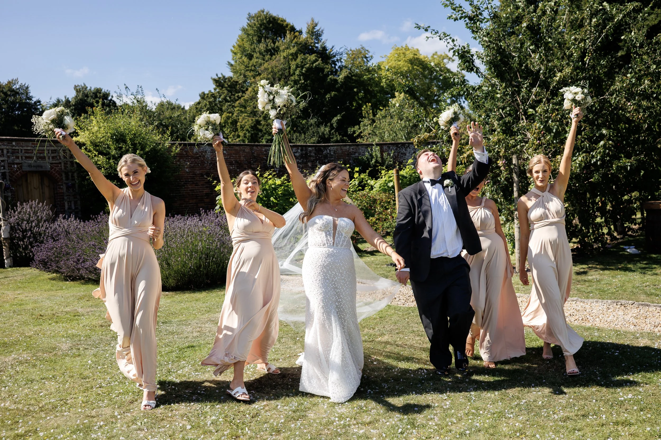 Bride and groom celebrating with bridesmaids outdoors on a sunny day, holding bouquets of white flowers, surrounded by trees and greenery.