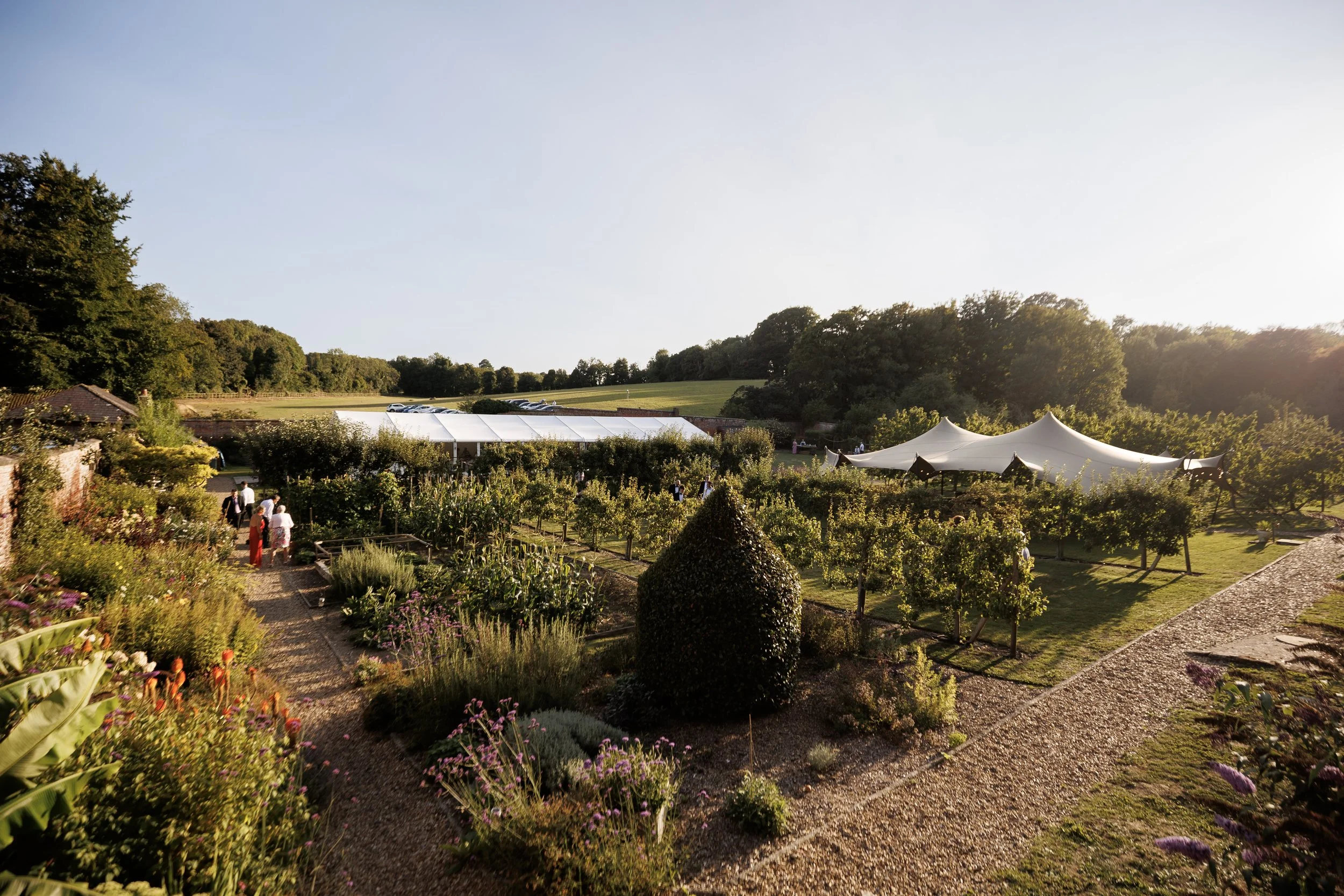 A lush garden with winding gravel paths, green bushes, and colorful flowers, with white tents set up on the grass and a few people walking and enjoying the outdoor setting in the evening sunlight.