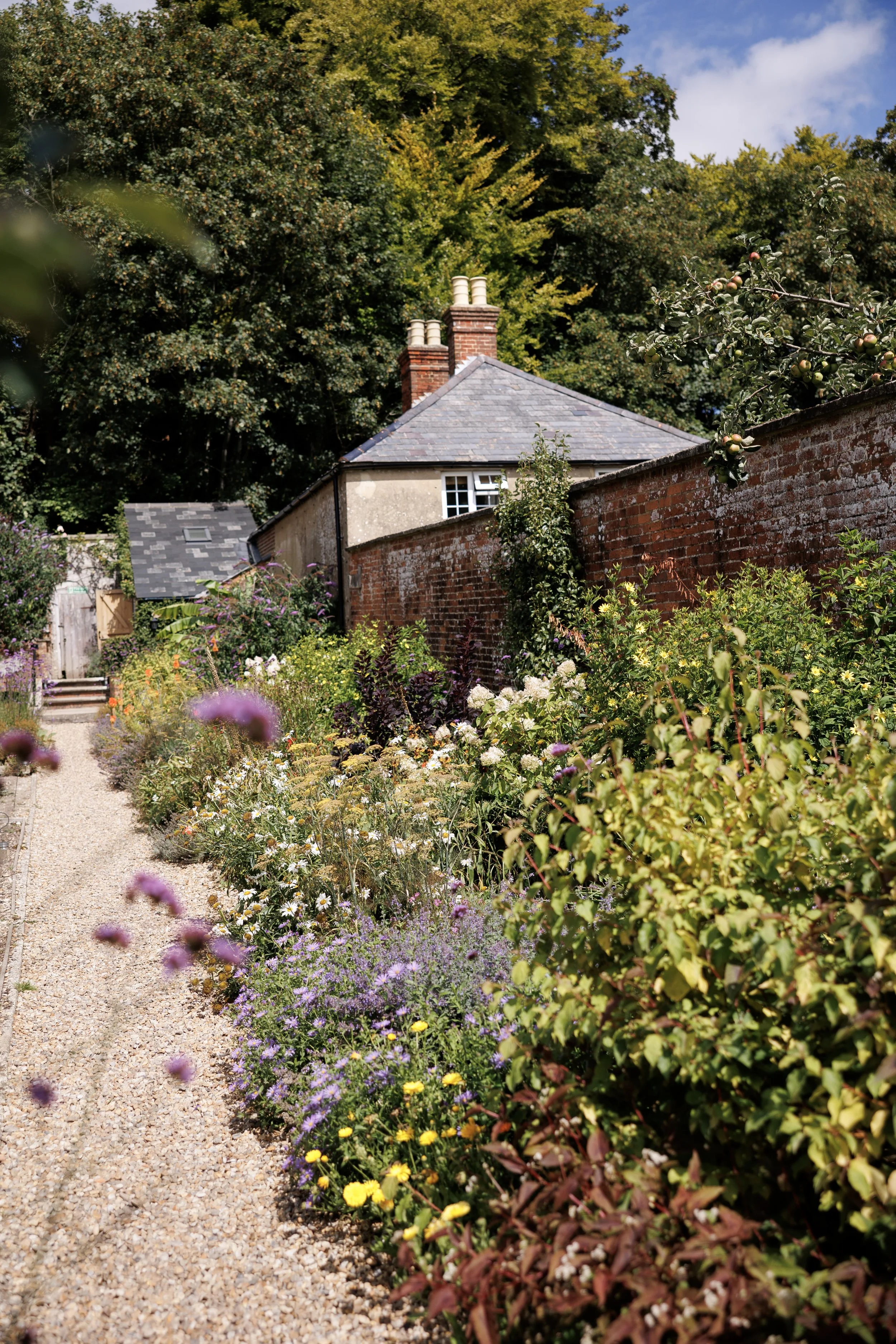 A lush garden with a gravel pathway flanked by colorful flowers and bushes, behind an old brick wall, and featuring a house with a slate roof and a chimney in the background, under a partly cloudy sky.