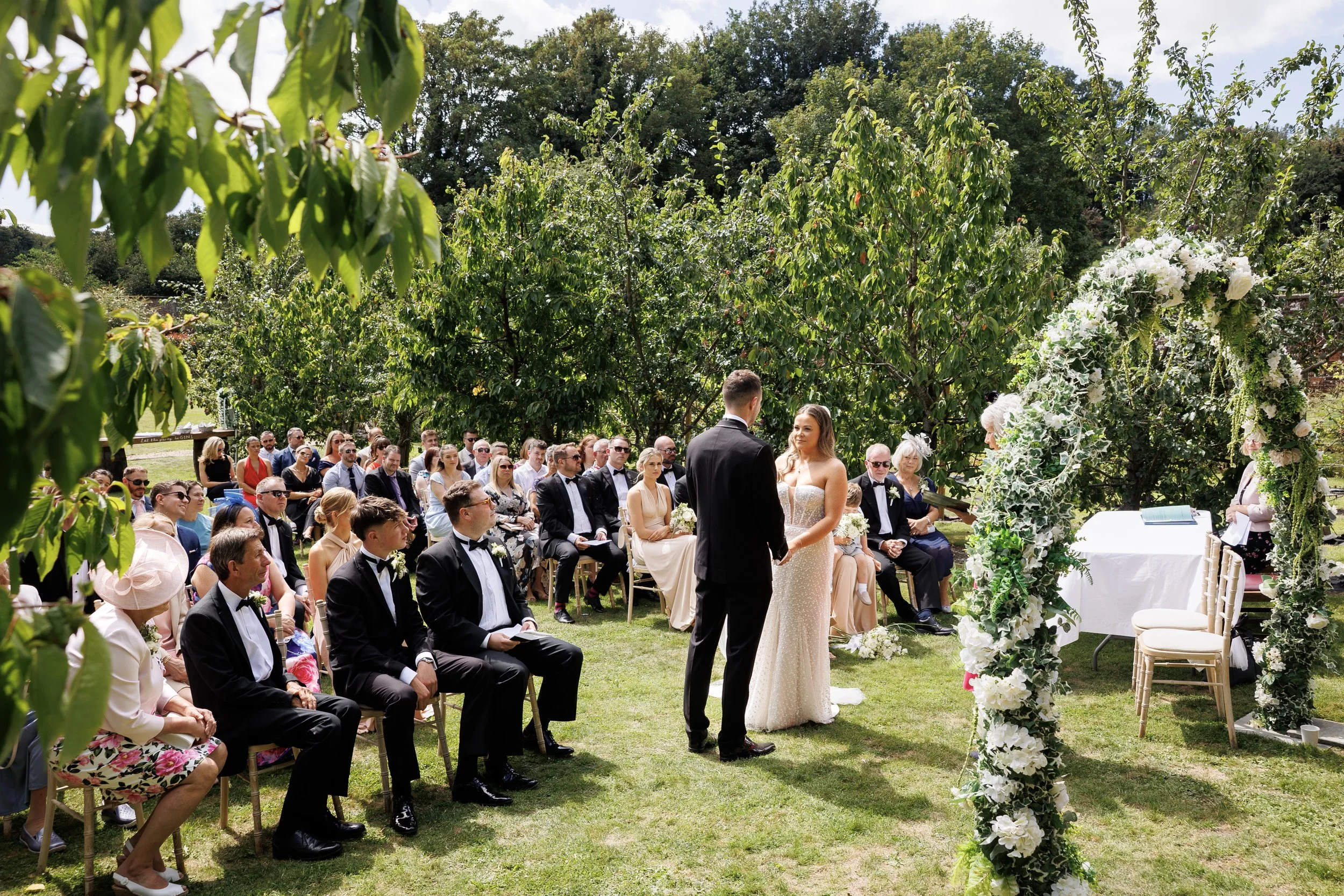 A couple gets married outdoors under a floral arch during a ceremony, with guests seated on grass and dressed in formal attire, surrounded by green trees.