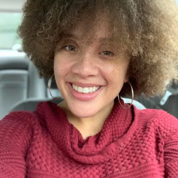 Young woman with curly hair smiling in a car, wearing hoop earrings and a red textured sweater.