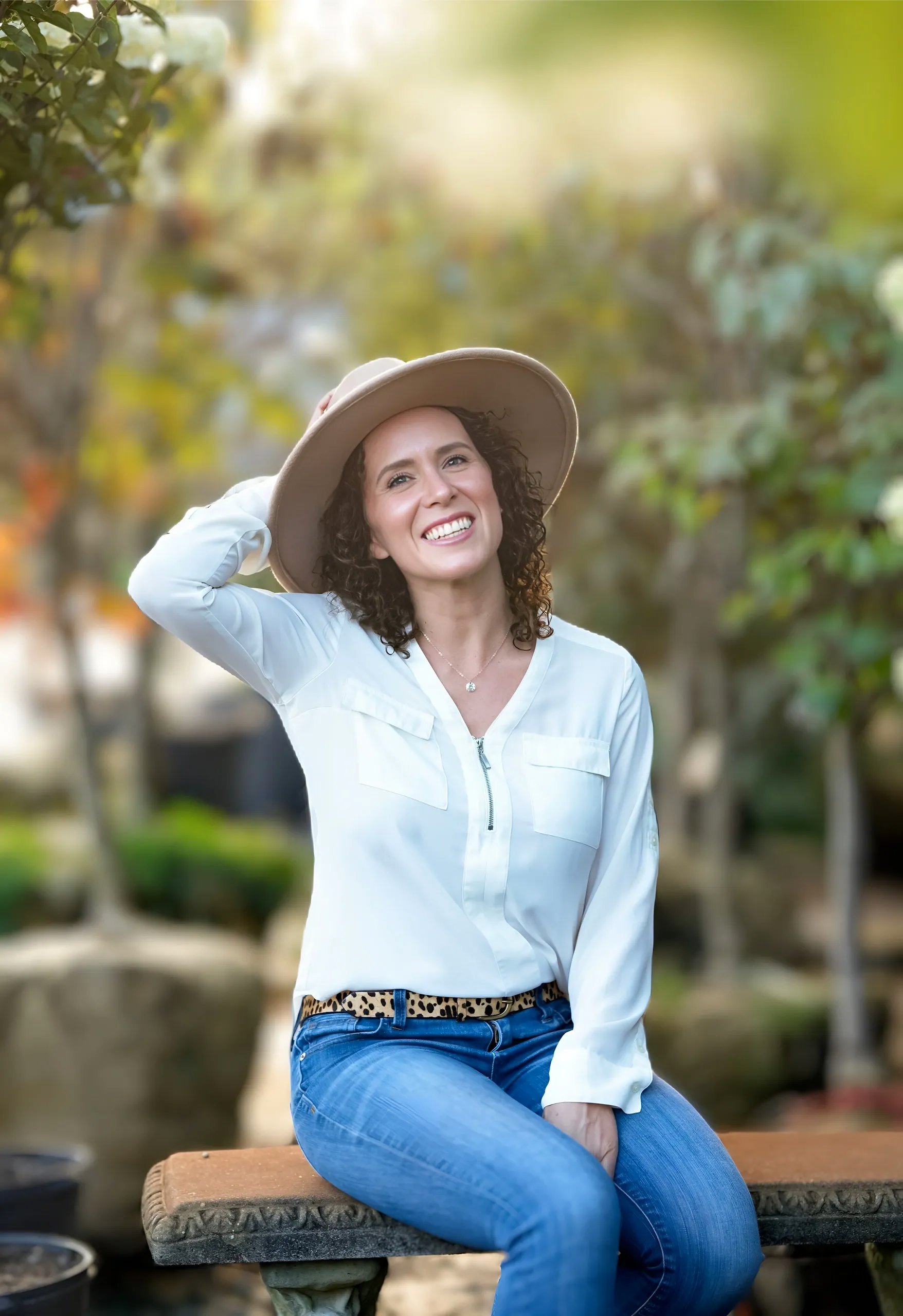 A woman sitting outdoors on a wooden bench, smiling, wearing a wide-brimmed hat, a white shirt, jeans, and a leopard-print belt, with greenery and blurred plants in the background.
