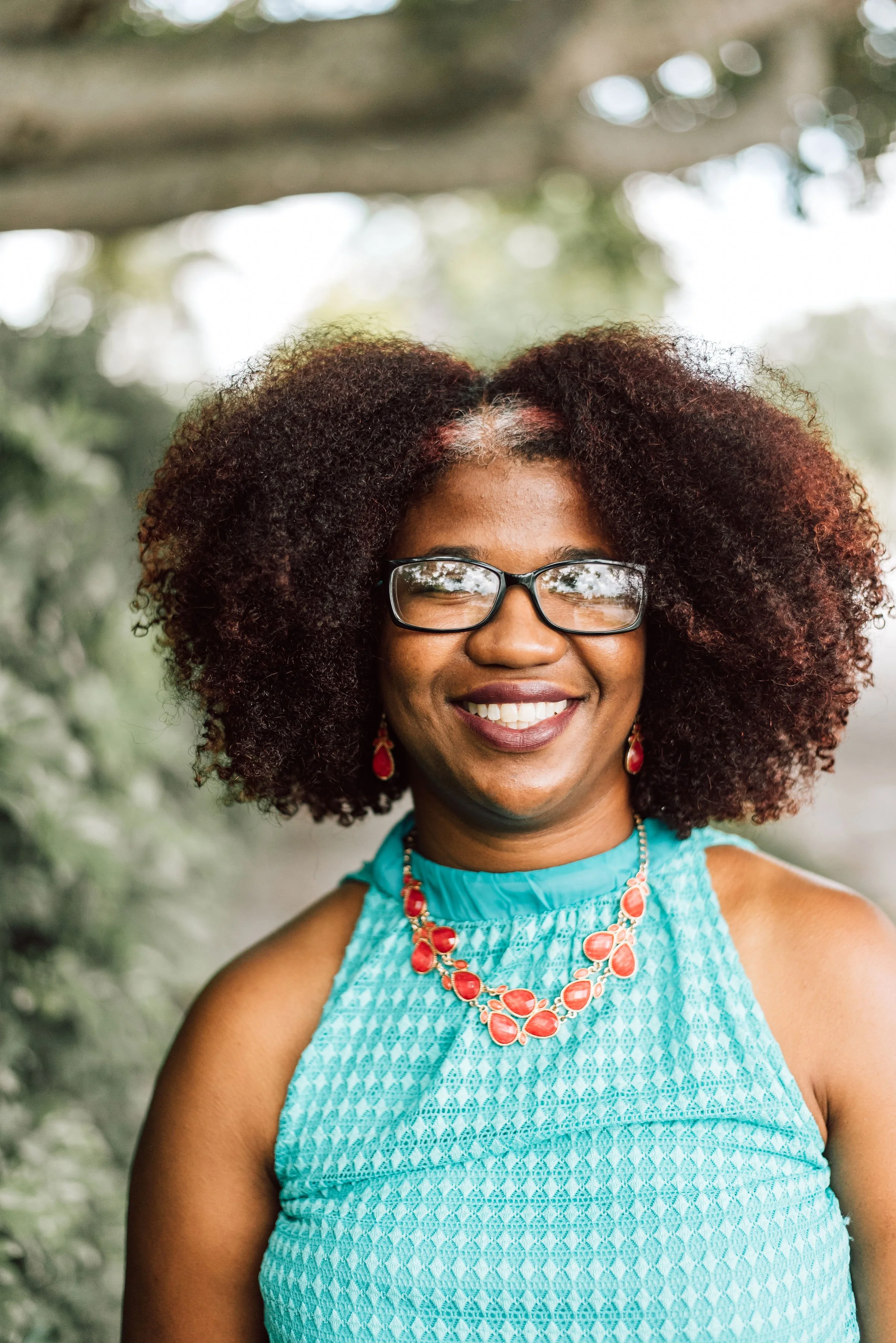 Smiling woman wearing glasses, a turquoise sleeveless top, and vibrant red jewelry outdoors with greenery in the background.