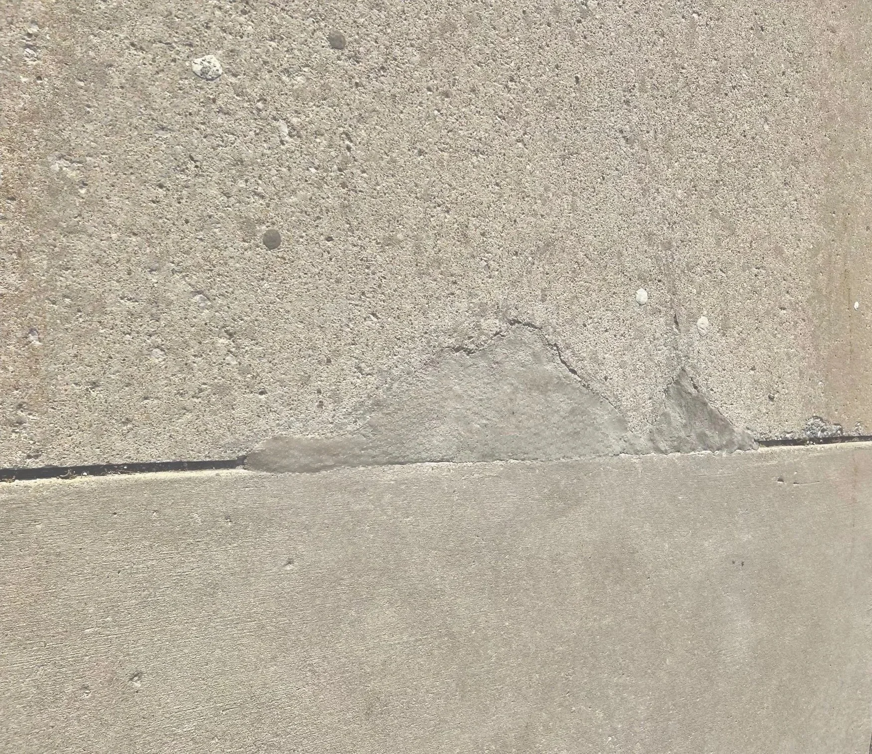 Close-up of a concrete sidewalk with cracks and pebbles, showing a line where two concrete slabs meet.