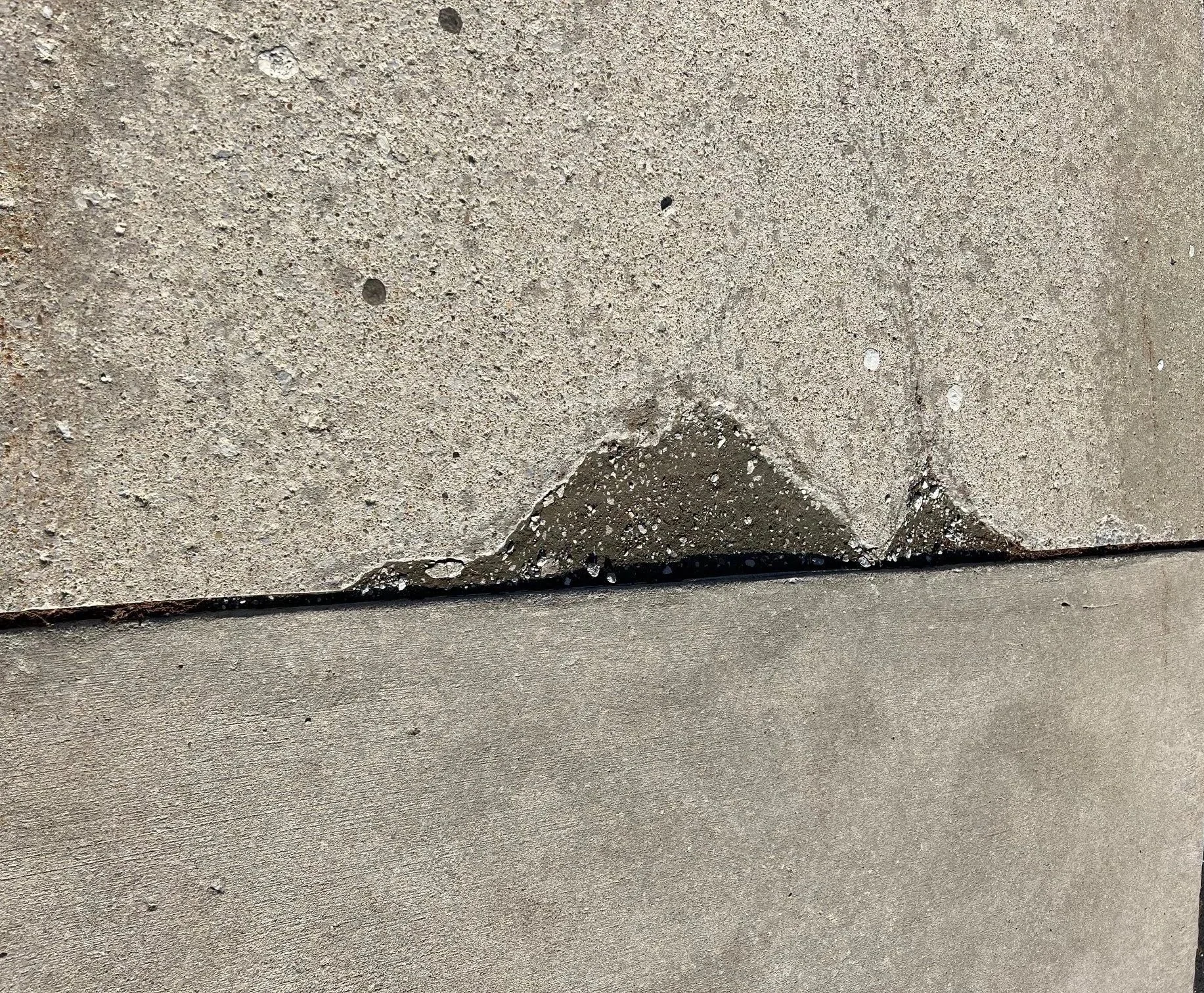 Close-up of concrete sidewalk with a concrete step and black rubber trim, showing some cracks and a small patch of repaired concrete.