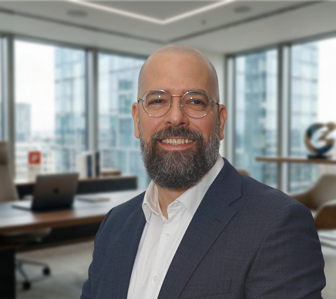 Sebastian Greschke trägt eine silberne Brille mit dünnem Rahmen, einen grau melierten Vollbart, ein weißes Hemd und einen dunkelblauen Anzug. Er lächelt. Hinter ihm sieht man einen unscharfen Büro-Hintergrund.