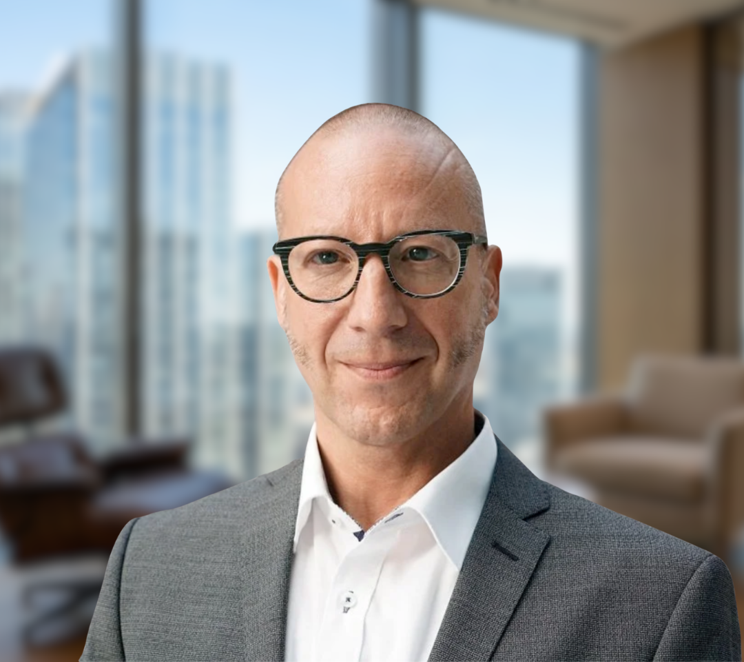 M. Gellner trägt eine dunkle, rundliche Brille, einen grauen Anzug und ein weißes Hemd vor einem hellen Büro-Hintergrund.