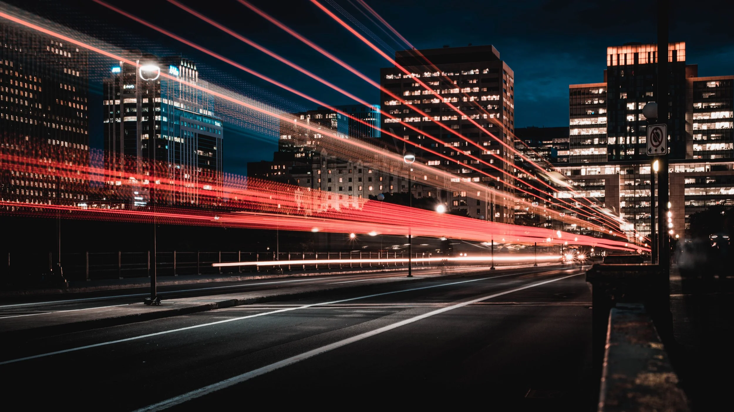 Nachtaufnahme einer Stadt mit beleuchteten Gebäuden und Lichtspuren von fahrenden Autos auf der Straße.