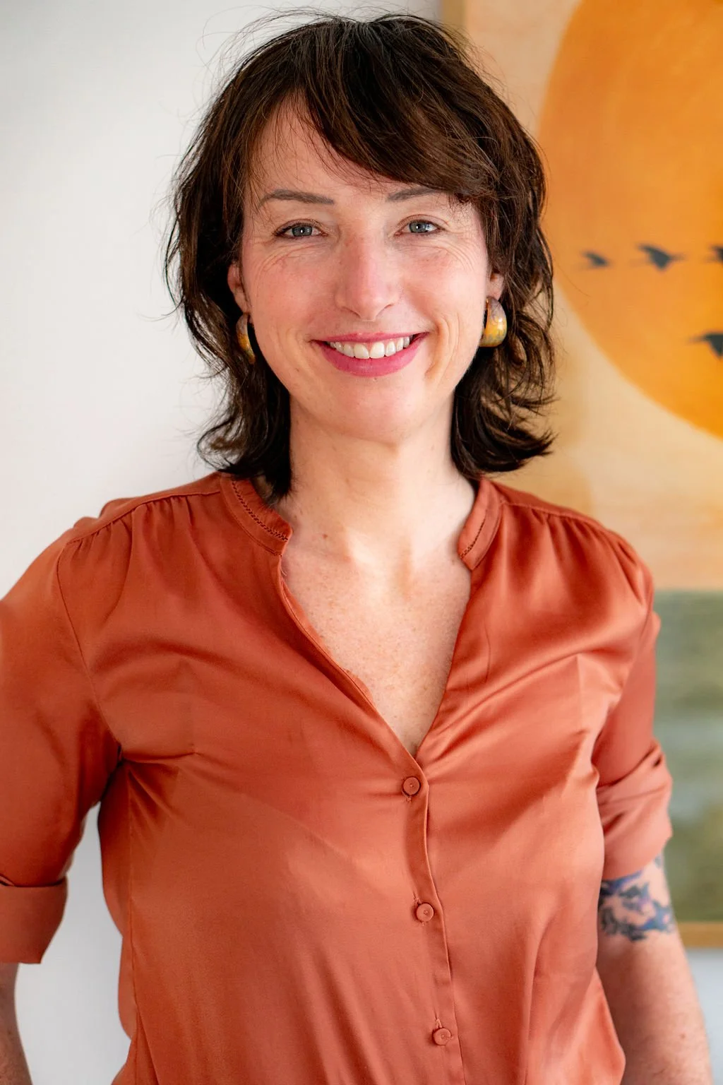 A woman with dark brown hair and blue eyes smiling, wearing an orange button-up shirt with rolled-up sleeves, standing indoors with a painting or print of a yellow circle and birds on a tan background in the background.