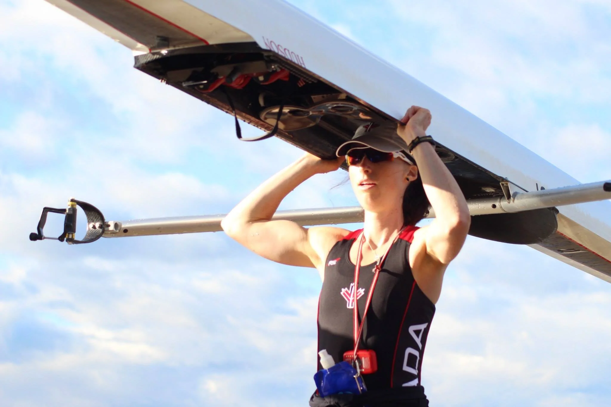 A female rower holding an indoor rowing shell on her shoulders, preparing for a training session outdoors on a partly cloudy day.