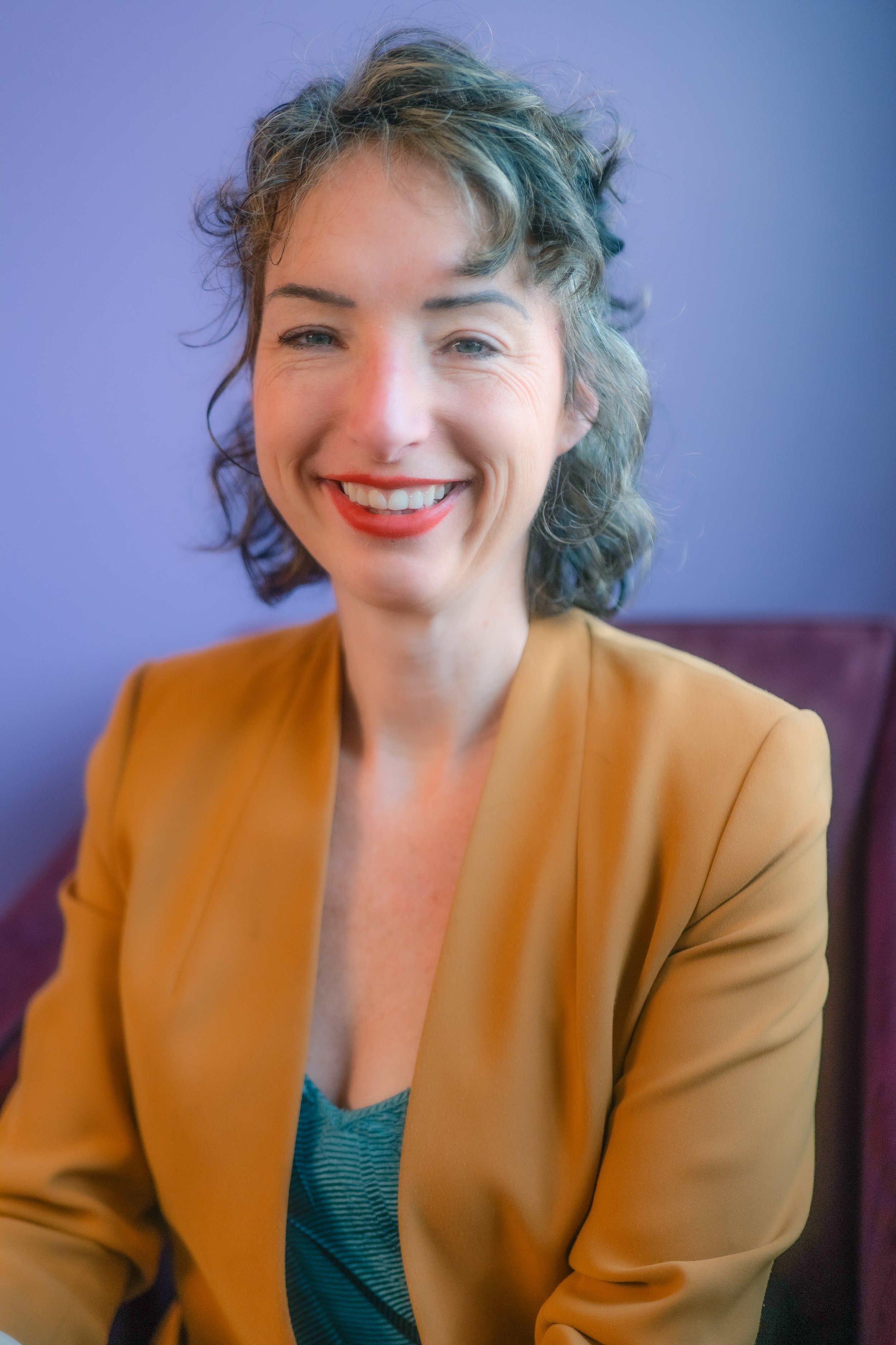 A woman with short, curly hair, smiling, wearing a mustard-colored blazer and a blue top, sitting against a purple wall.