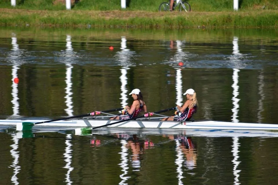 Two women in a racing boat on a calm body of water, wearing athletic clothing and white caps, with a grassy bank and a cyclist in the background.