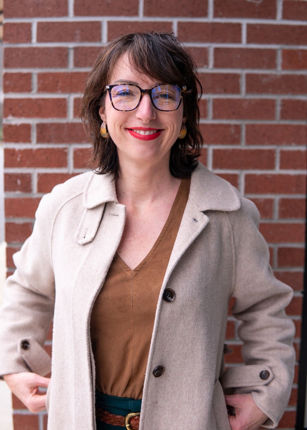 A woman with shoulder-length brown hair, wearing glasses, a beige coat, and a brown top, standing in front of a brick wall, smiling.