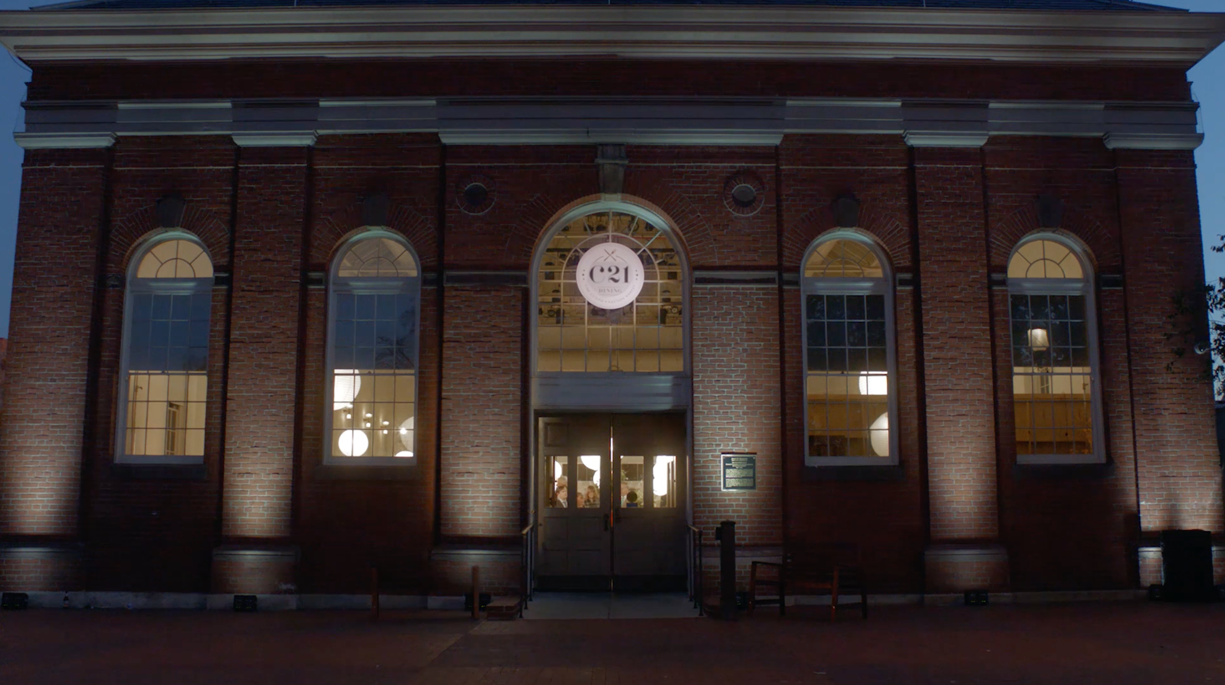 The exterior of a red brick building at night, with large arched windows and a circular sign above the door that reads 'C21 Design'. The building is illuminated from inside, revealing interior lighting and some furniture visible through the windows.