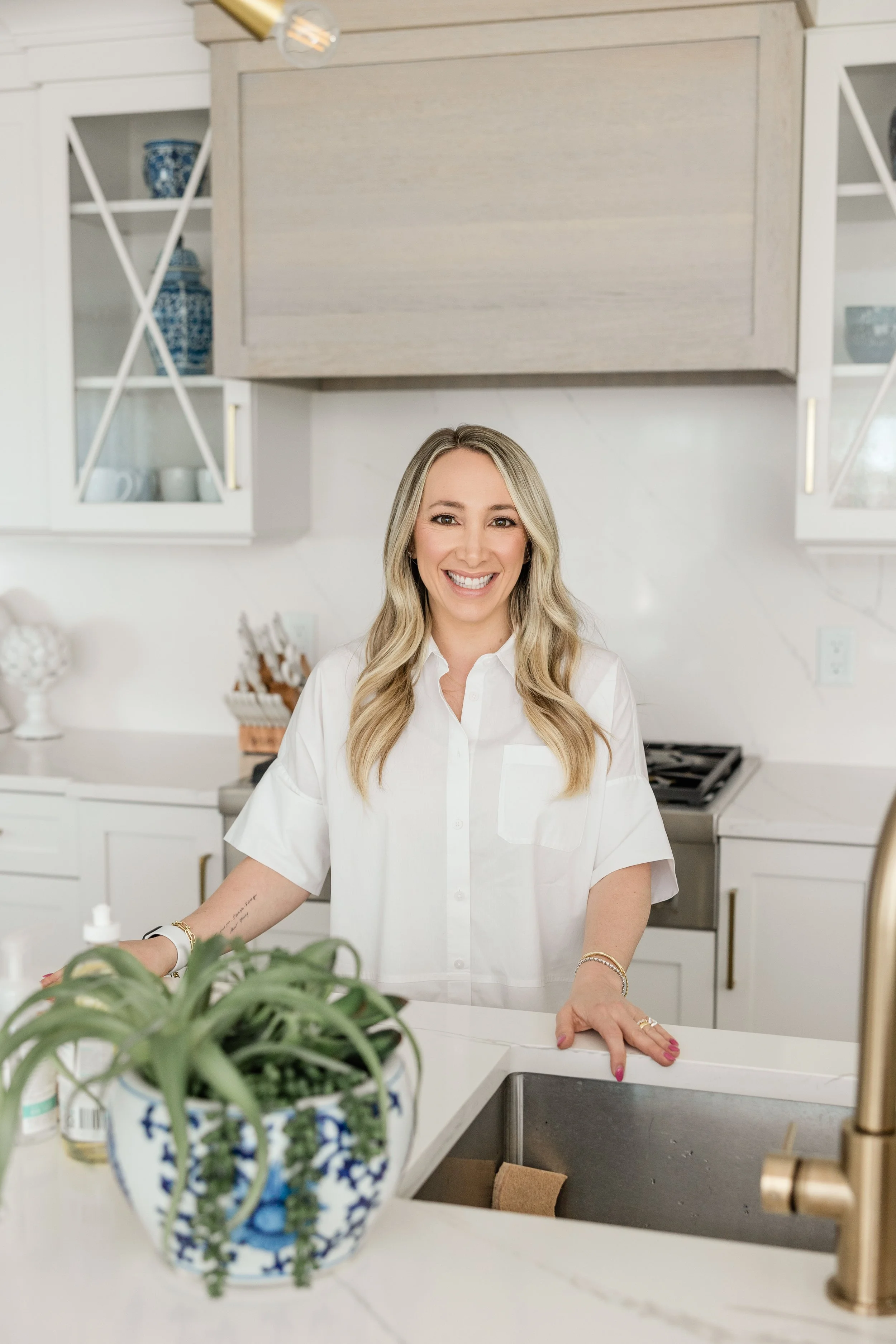 A woman with blonde hair smiling while standing in a modern kitchen, wearing a white shirt, with decorative items and cabinets in the background, and plants in the foreground.