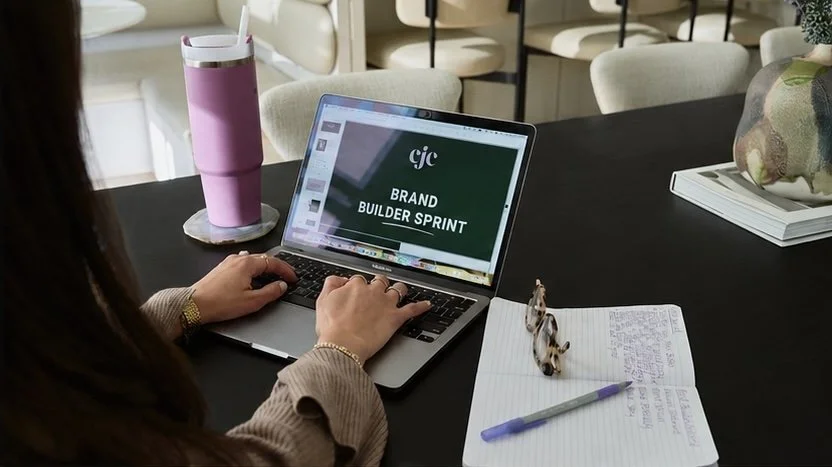 A woman working at a black table with a laptop displaying a presentation titled "Brand Builder Sprint." On the table, there is a pink tumbler, a pair of sunglasses, a purple pen, and a notebook with handwritten notes. In the background, there are chairs and a decorative vase.