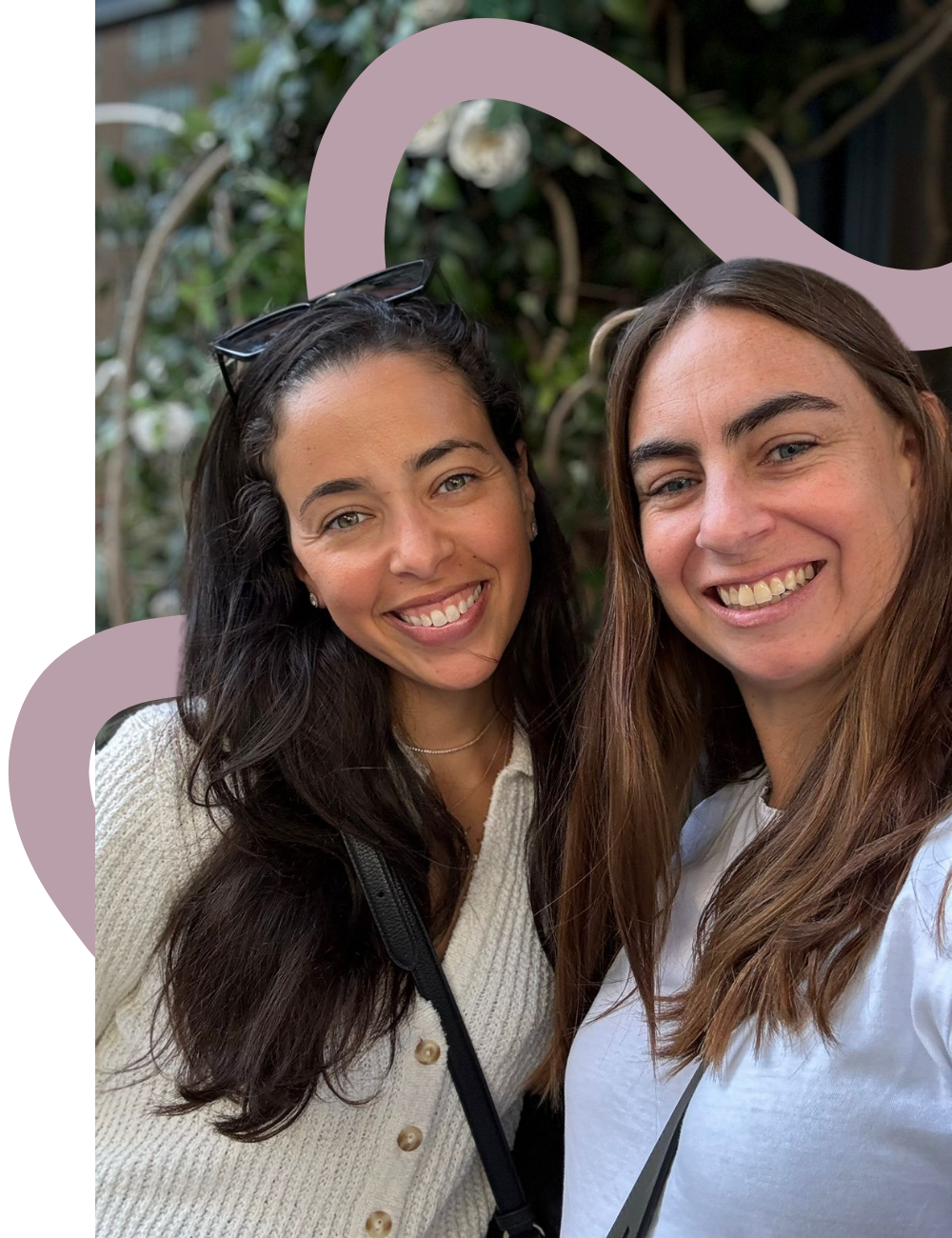 Two women smiling and taking a selfie outdoors, with greenery and a building in the background.