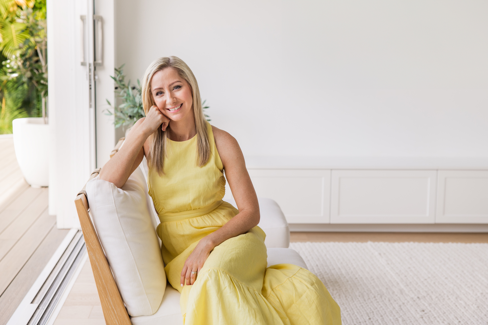 A smiling woman with blonde hair in a yellow dress sitting on a white sofa in a bright room with large windows and greenery outside.