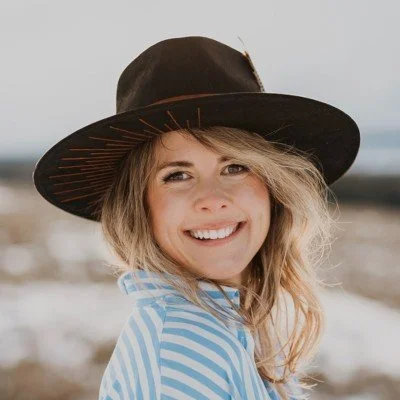 A smiling woman with wavy blonde hair wearing a wide-brimmed black hat and a blue and white striped shirt, standing outdoors in a natural setting.