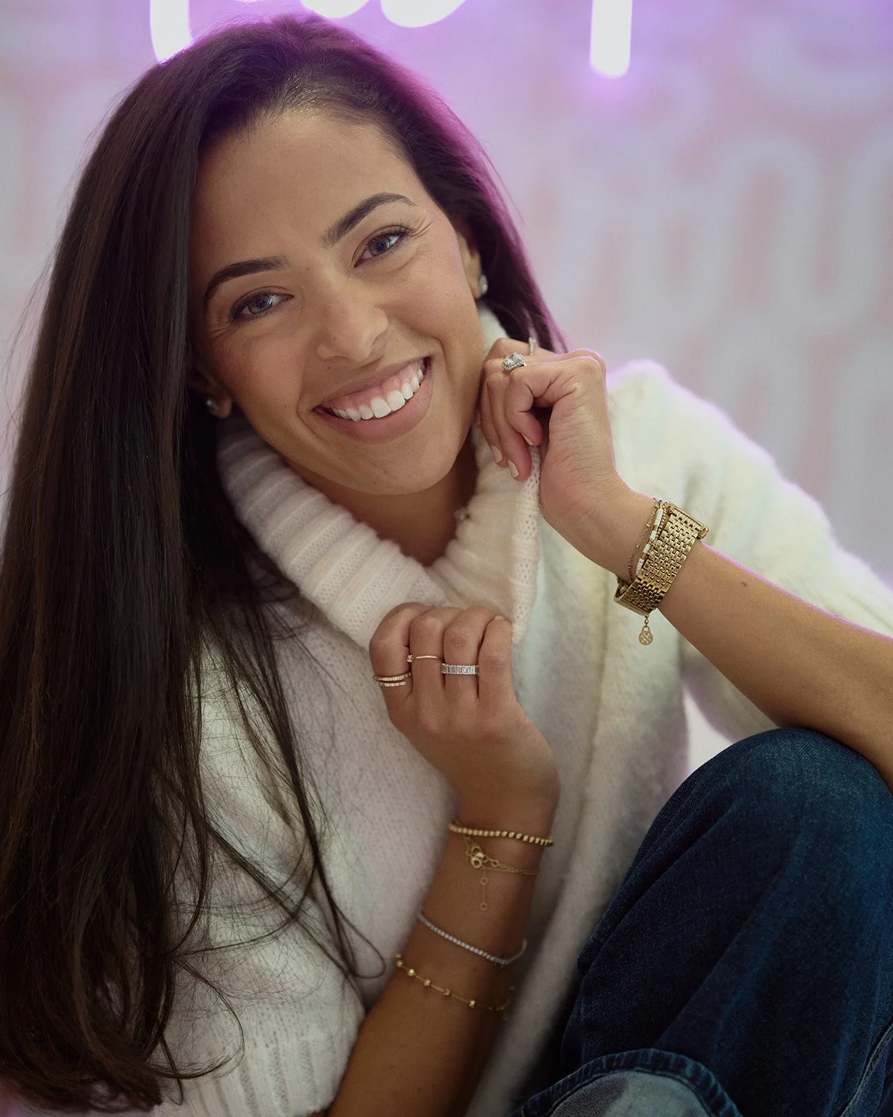 A woman with long dark hair smiling, wearing a white turtleneck sweater, gold jewelry, and sitting in front of a purple and pink background.