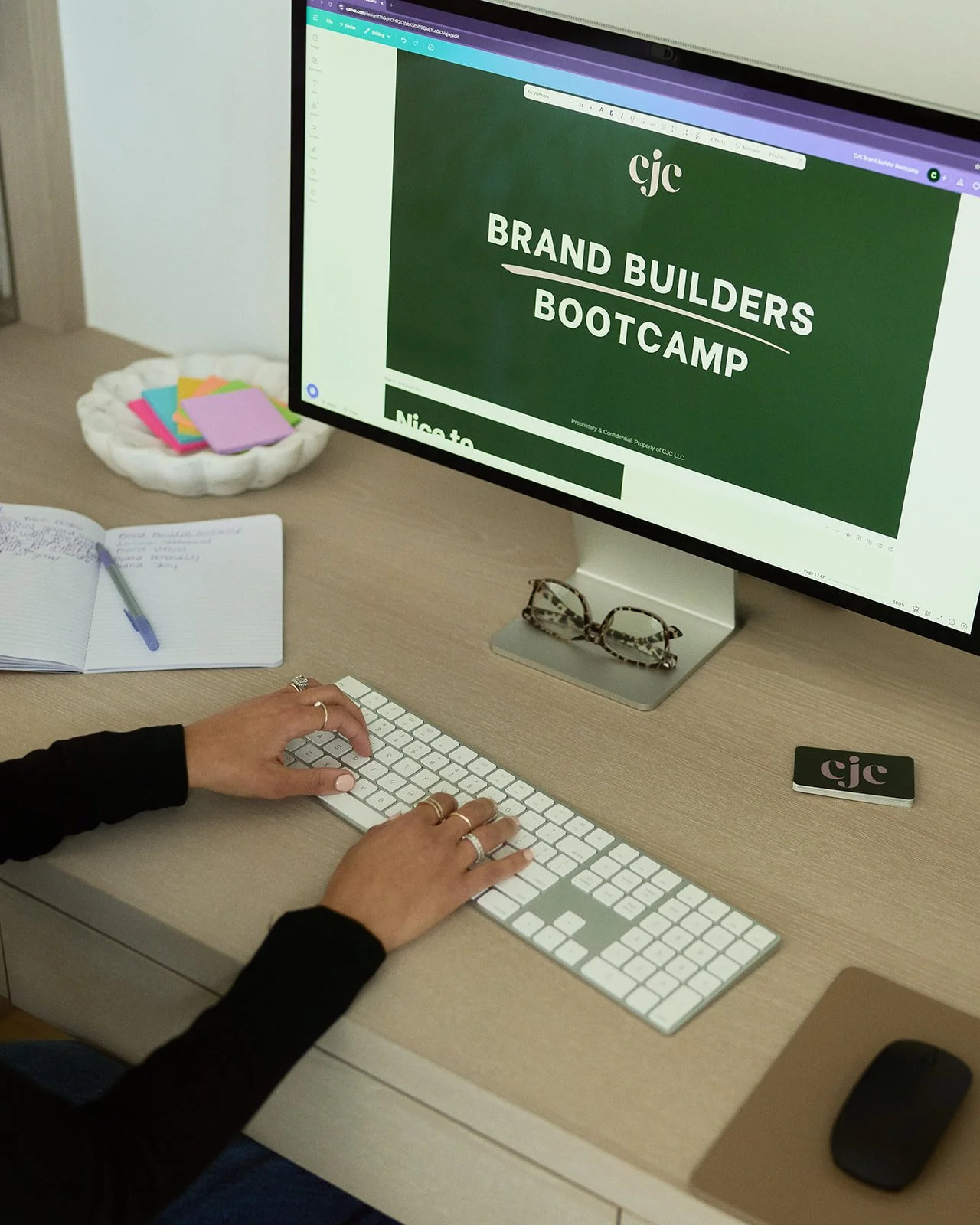 A person using a computer with a large monitor displaying a presentation titled 'Brand Builders Bootcamp.' The person is typing on a white keyboard, wearing rings on their fingers. Nearby on the desk, there are a pair of glasses, a closed smartphone, and an open notebook with a pen. To the left, there is a dish with multicolored sticky notes, and an open notebook with handwritten notes.