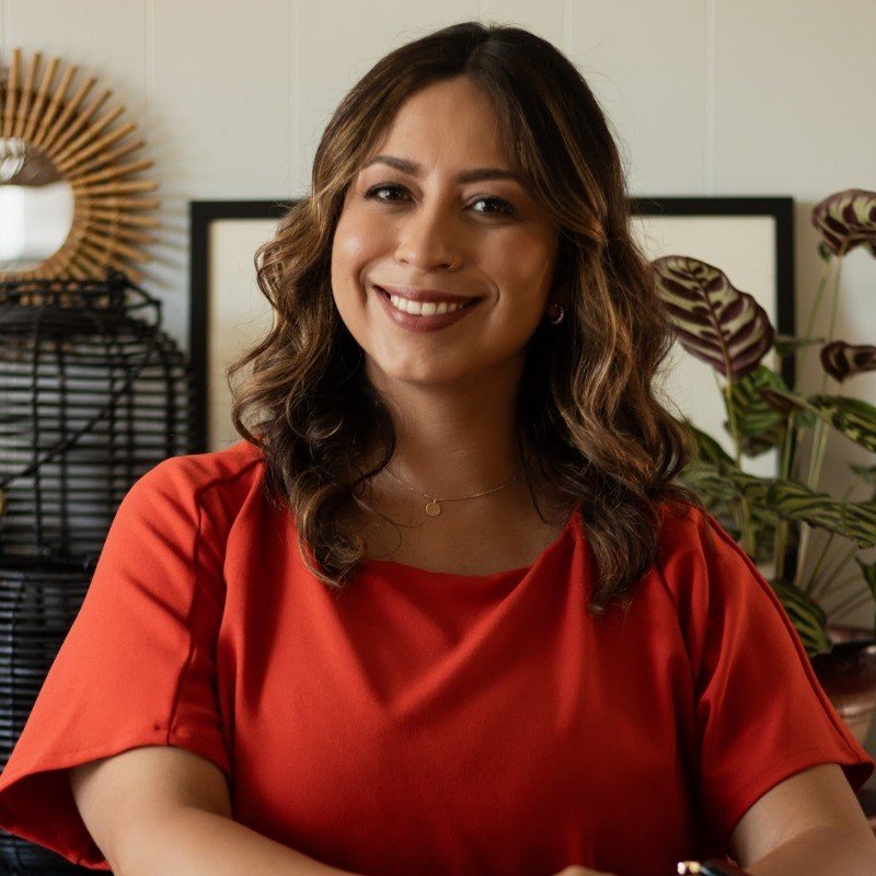 A smiling woman with wavy brown hair, wearing a red top, sitting indoors with a background of houseplants and decorative items.